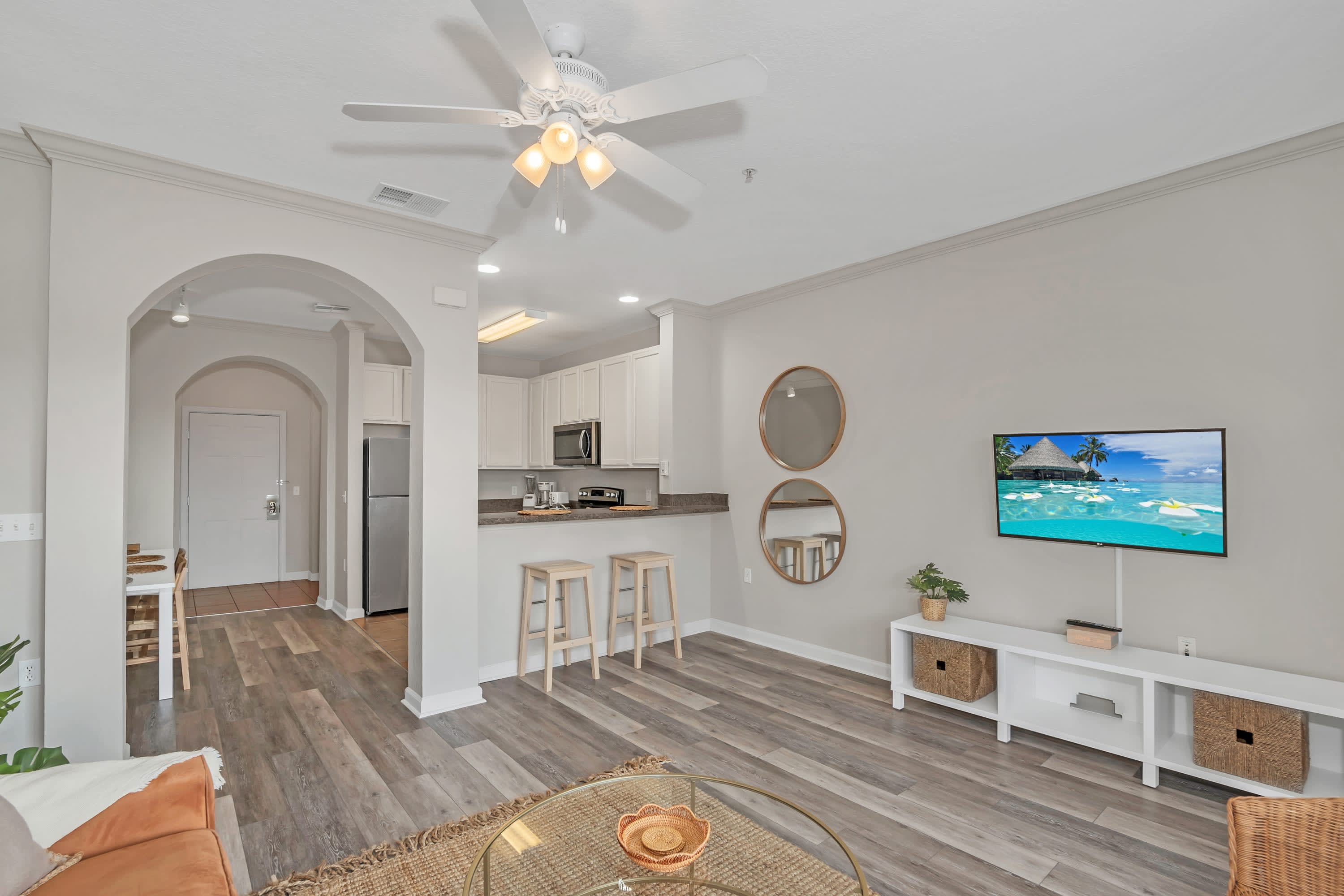 A wide shot of the living room showing a wall-mounted TV, a white media console, and a view into the kitchen with a breakfast bar and two stools. The room has wood-look flooring and a ceiling fan.
