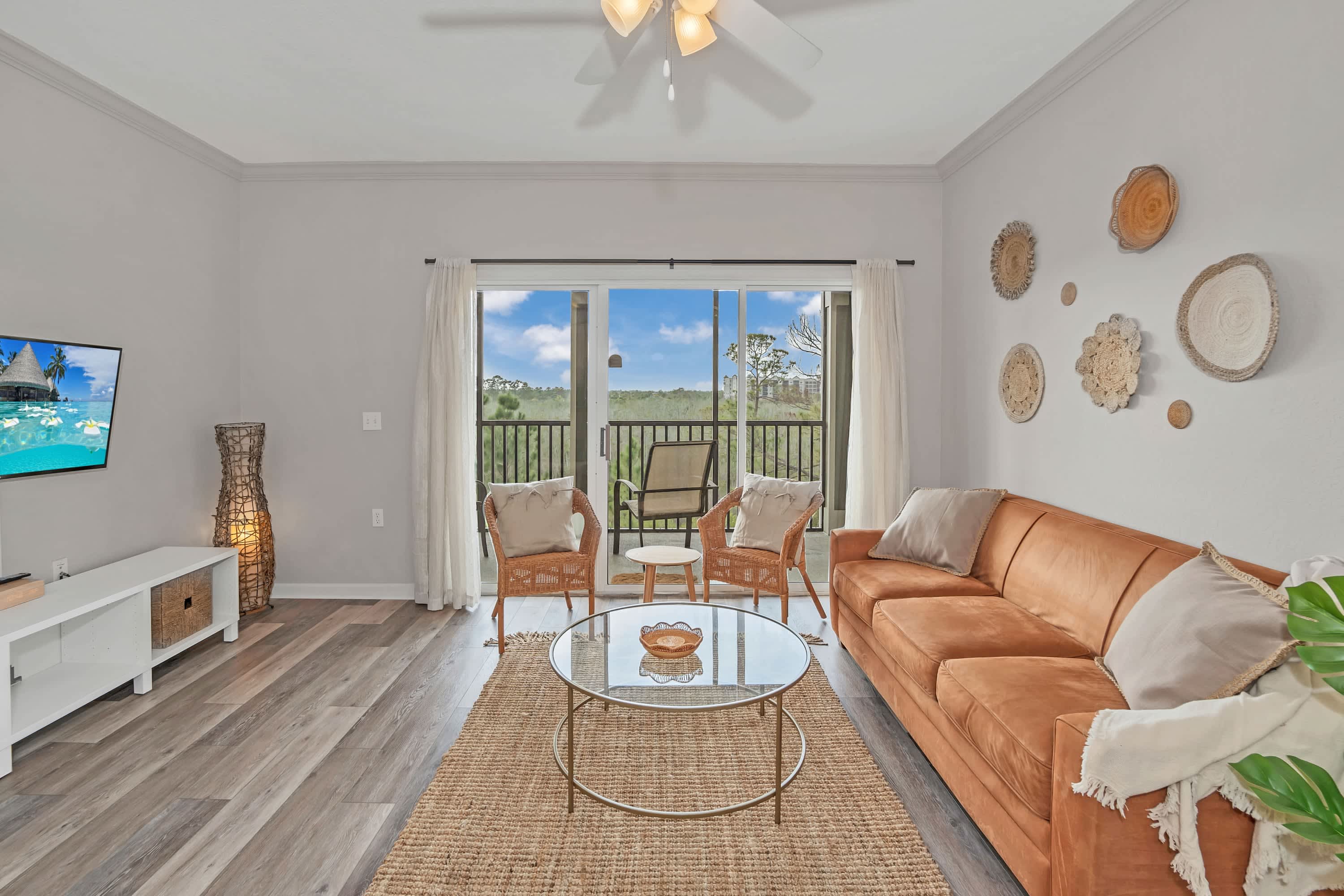 A bright living room with a large orange sofa, a round glass coffee table on a textured jute rug, and decorative woven wall art. Sliding glass doors lead to a balcony with outdoor seating and a view of trees.