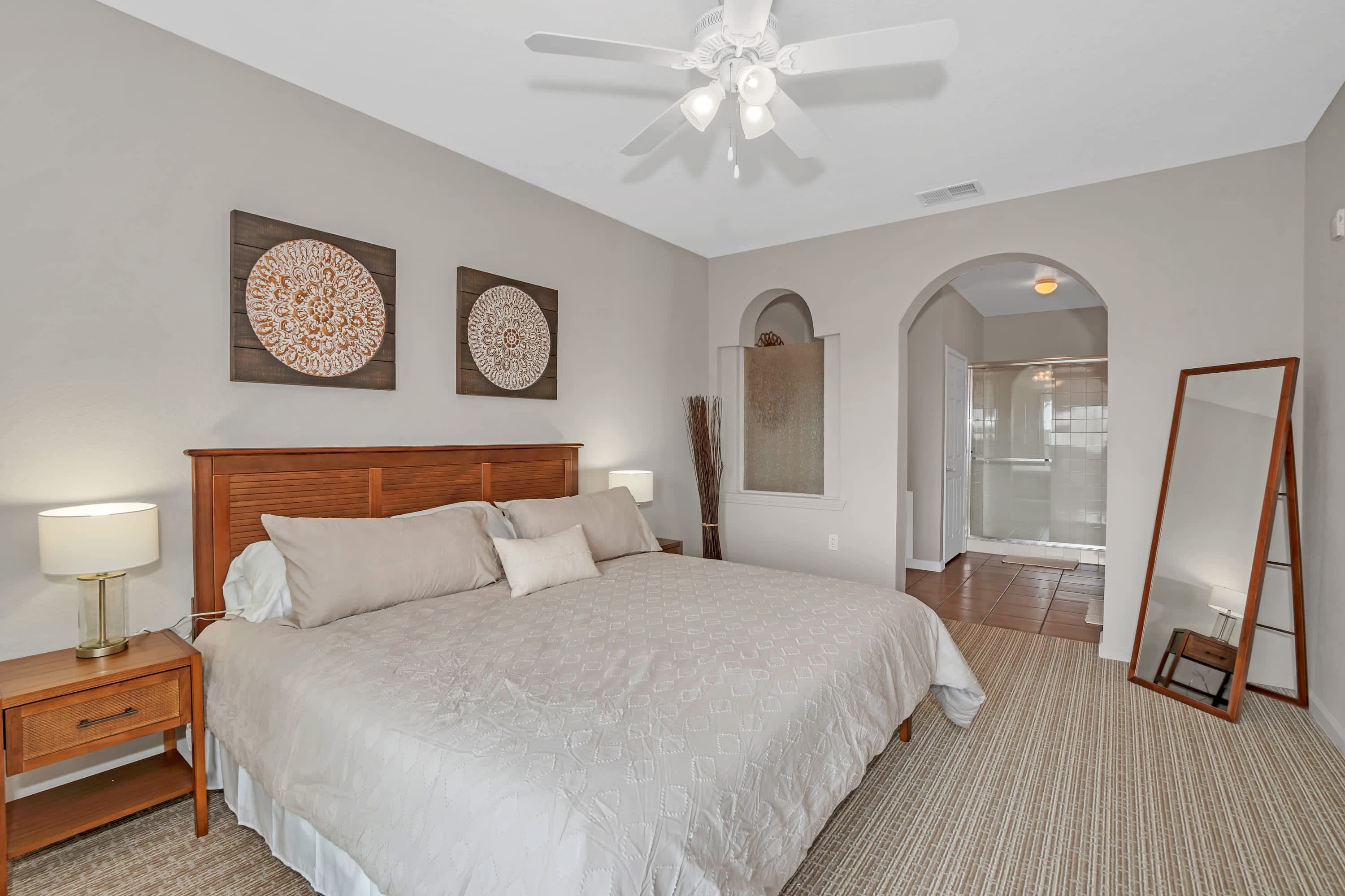 A spacious master bedroom featuring a king-sized bed with a wooden headboard, light-colored bedding, and two circular decorative wall hangings above the bed. Arched doorways lead to an en-suite bathroom with a shower visible.