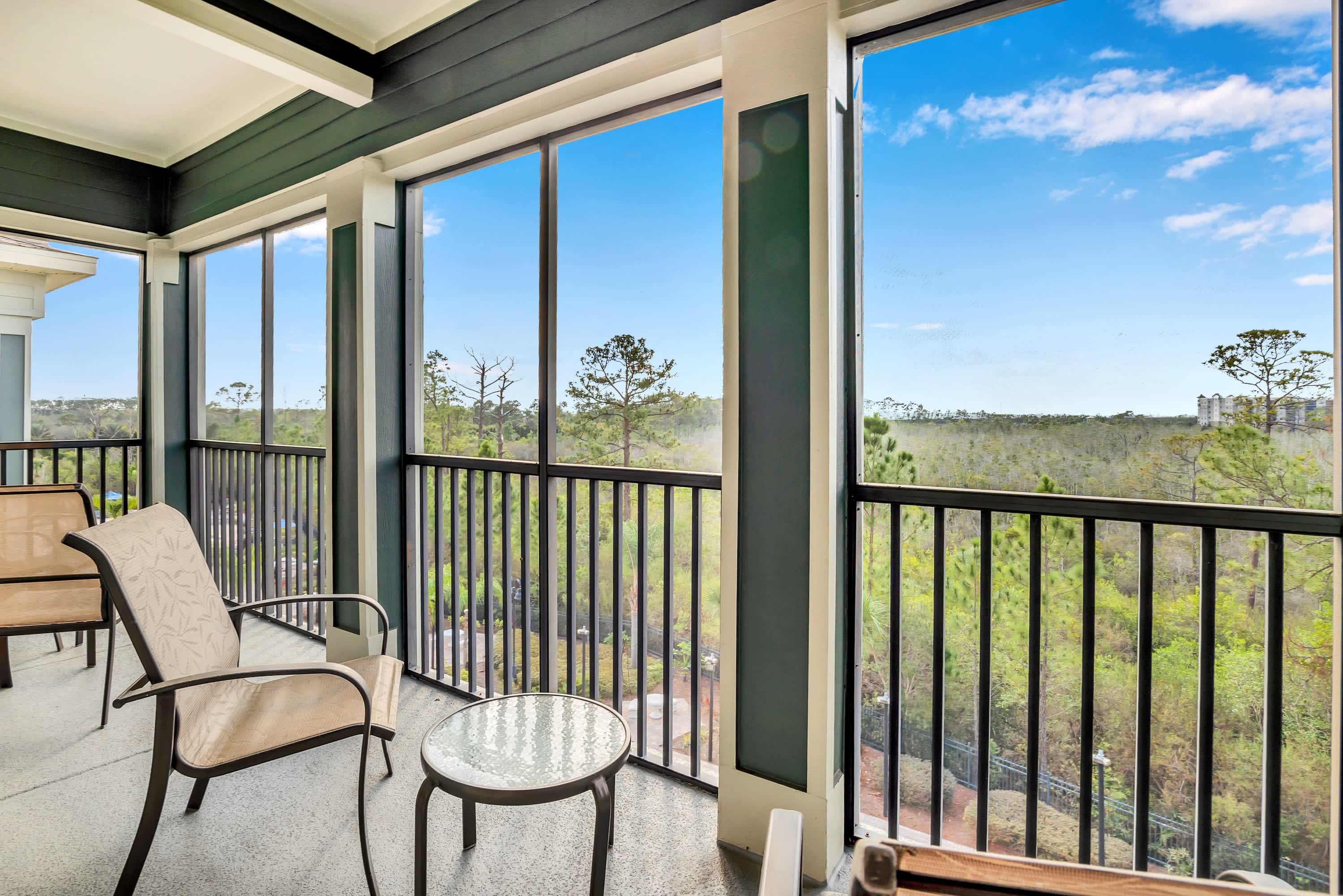 A screened-in balcony with dark railings and pillars, offering a panoramic view of a lush green landscape with trees and distant buildings under a bright blue sky. There's outdoor seating with a small glass-top table and chairs.