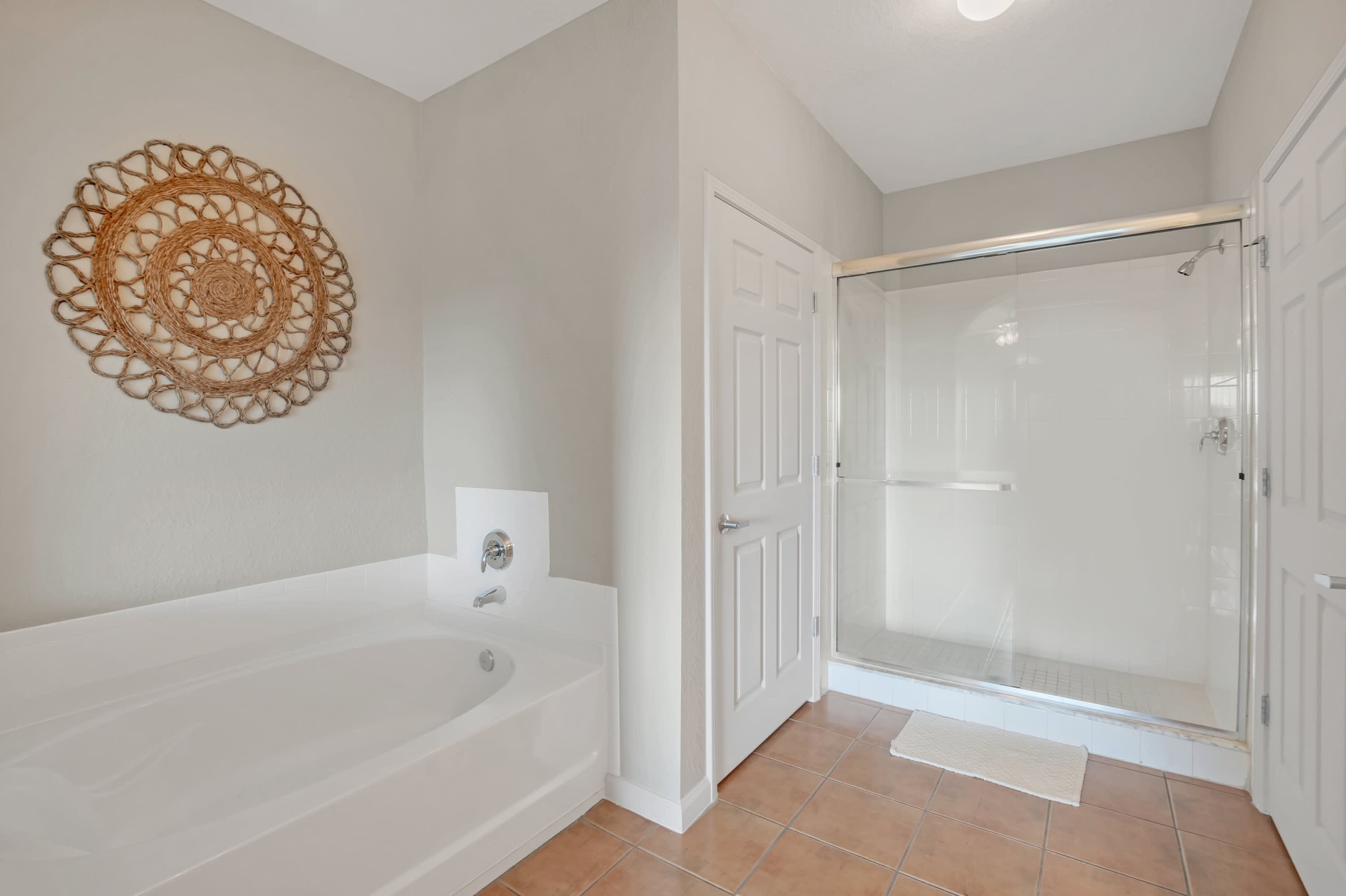 Another view of the master bathroom, showcasing a large white soaking tub and a separate glass-enclosed walk-in shower. The floor is tiled in a terracotta color, and a woven circular wall decoration hangs on the wall.