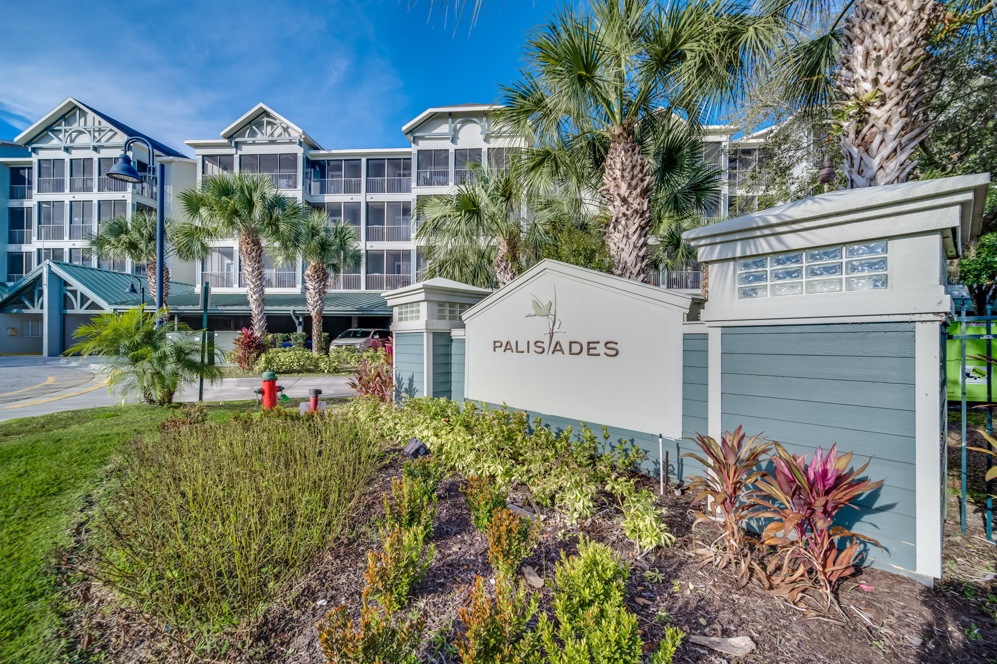 The entrance sign for 'PALISADES' stands prominently amidst lush green landscaping, with a multi-story condominium building featuring screened balconies and palm trees under a bright blue sky in the background.