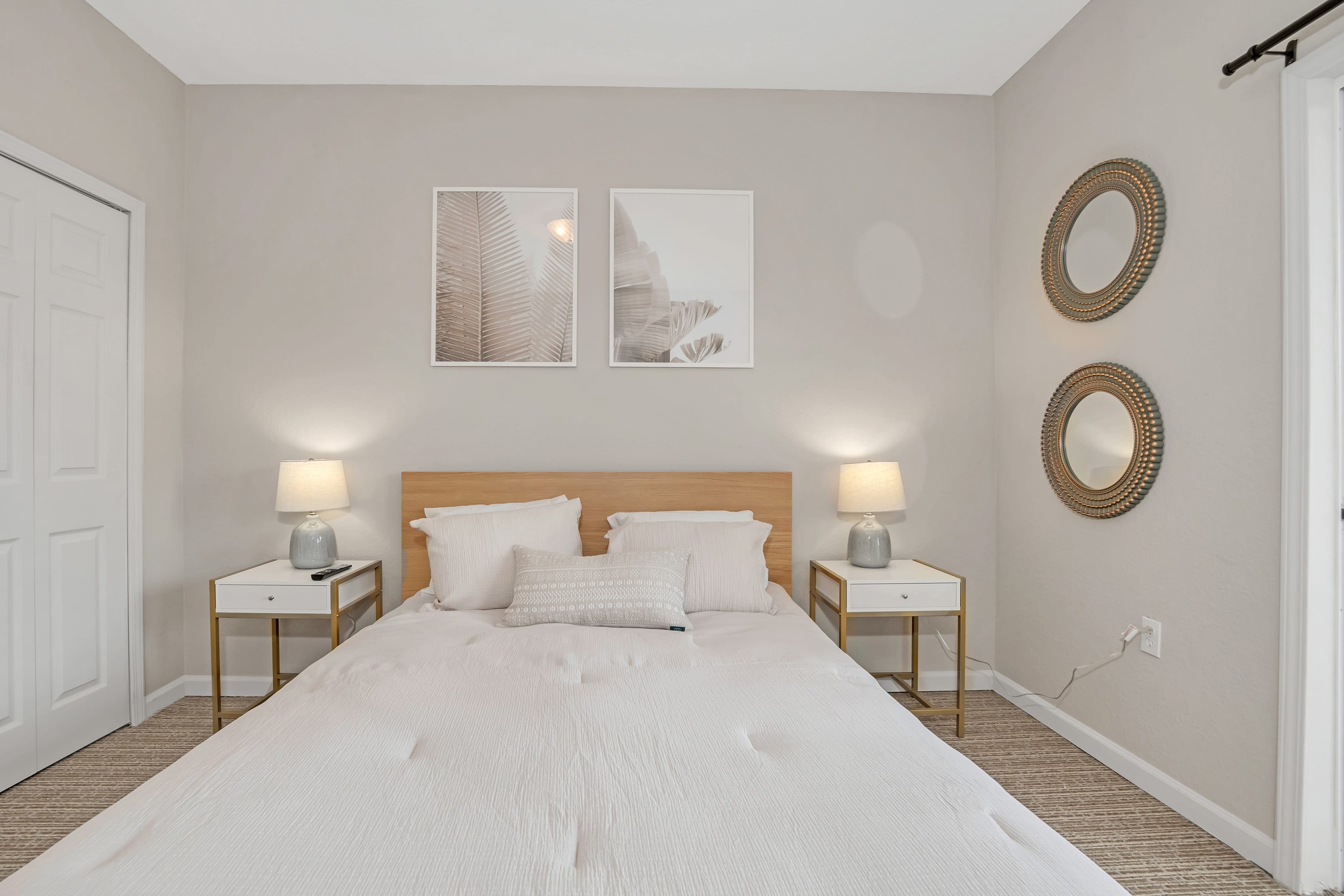 A closer view of the queen-sized bed in bedroom 2, showing the light wood bed frame, neutral bedding, and two white nightstands with lamps. Two framed botanical prints hang above the bed, and two round mirrors are on the adjacent wall.