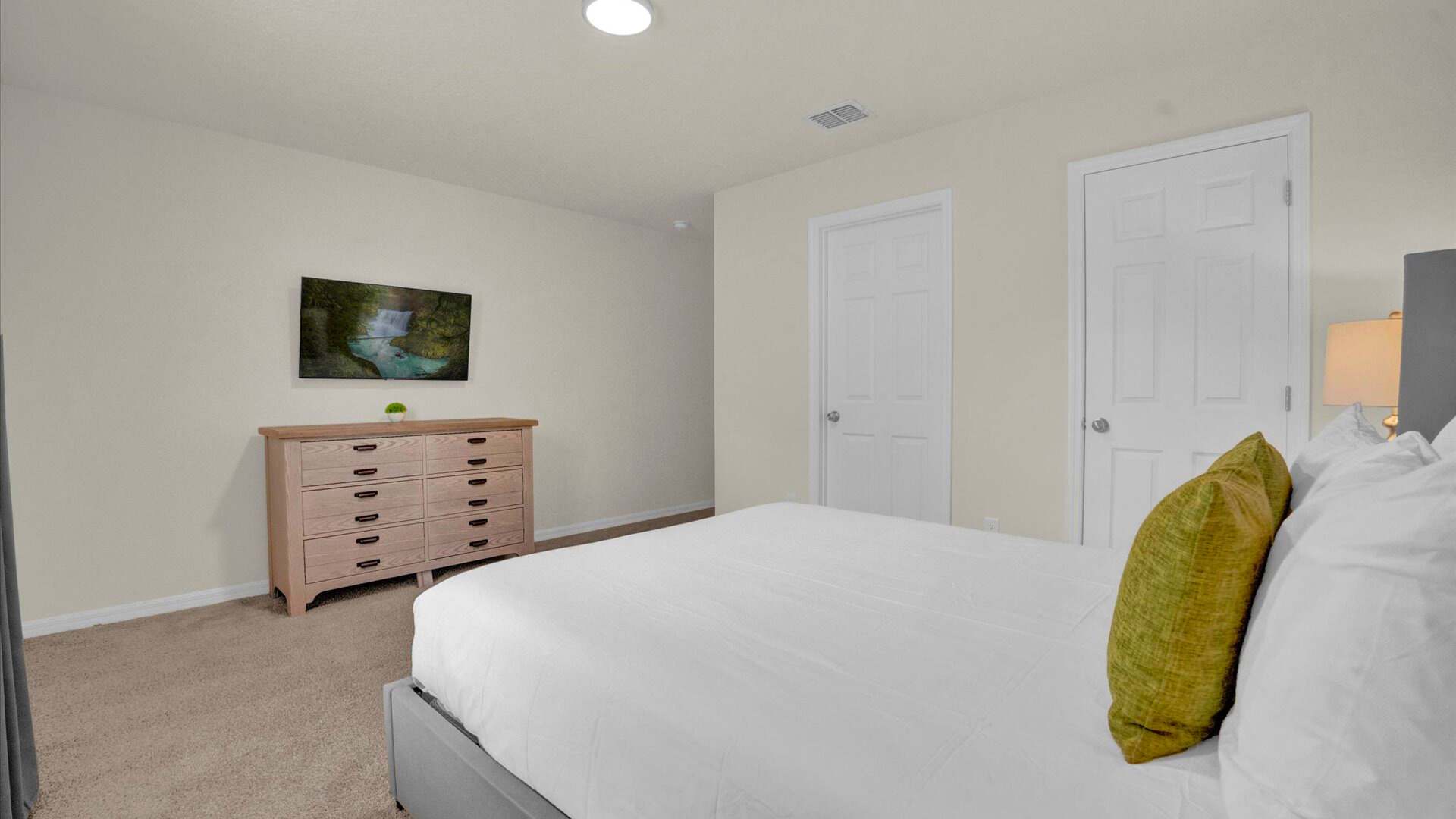 View of the master bedroom showing a light wooden dresser with a wall-mounted TV above it. The room has light beige carpet, a tray ceiling with recessed lighting, and two white doors, likely leading to a closet and bathroom.