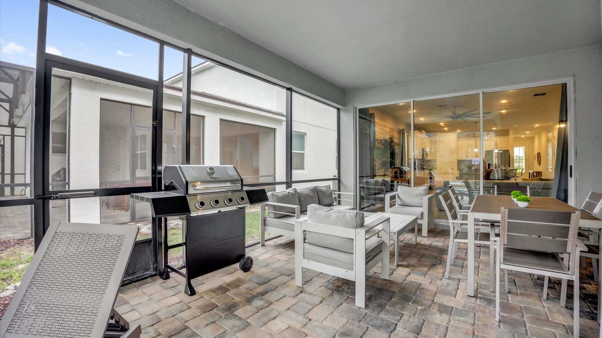 Wide view of a covered outdoor patio featuring a gas BBQ grill, a white outdoor seating arrangement with a sofa and armchairs, and a dining table, all situated on brick pavers within a screened area.