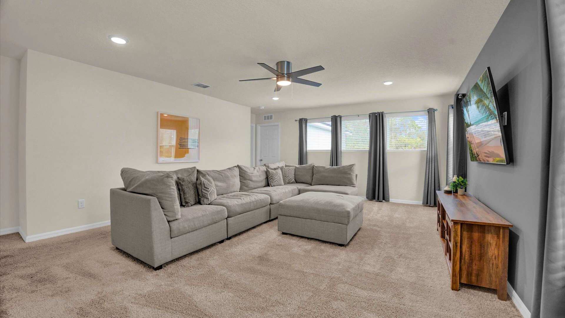 Spacious living room with light beige carpet, a large L-shaped grey sectional sofa with an ottoman, and a ceiling fan. A wall-mounted TV is visible on a dark accent wall, flanked by windows with blinds and grey curtains.