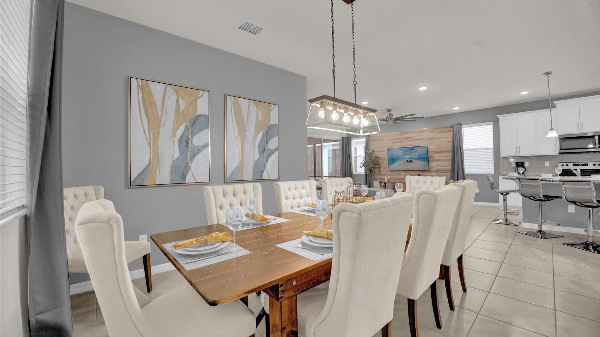Stylish dining room with a long wooden table and eight upholstered chairs, illuminated by a contemporary linear chandelier. Abstract artwork adorns the light gray wall, complementing the open-concept layout that connects to the living area.