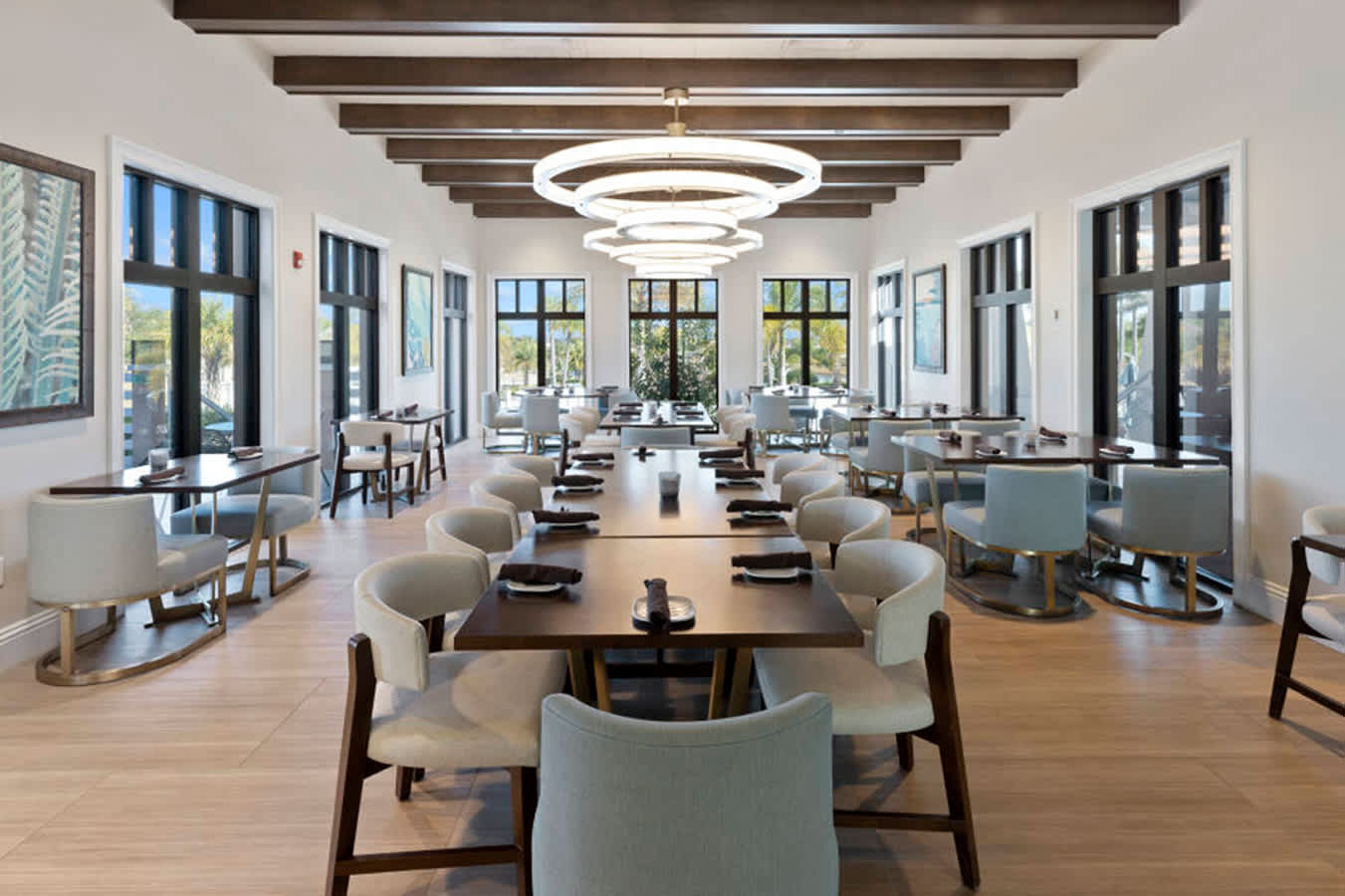 A spacious, modern dining room features multiple long dark wood tables with light upholstered chairs, set on light wood-look flooring. Overhead, a series of large, multi-ringed circular chandeliers illuminate the space, which is framed by dark-beamed ceilings and tall windows.