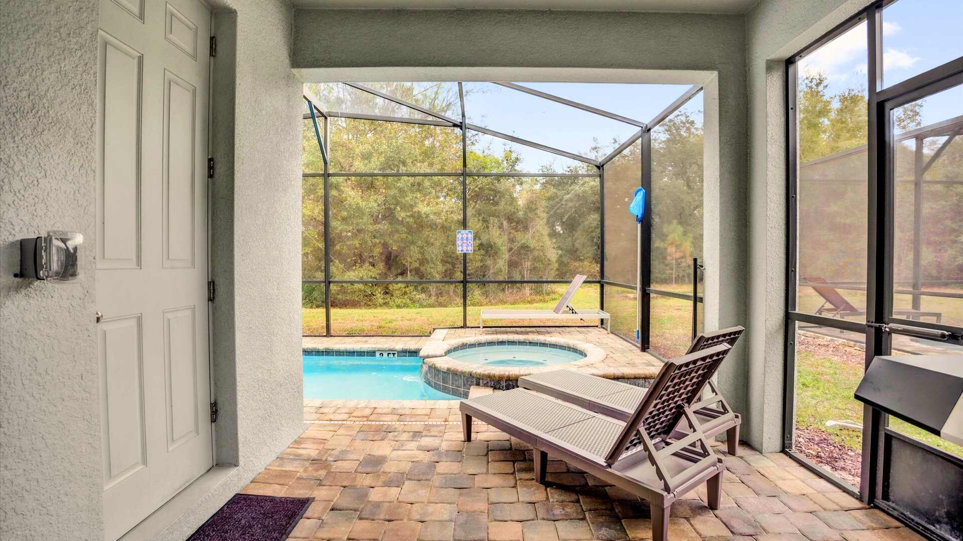 Covered patio area with direct access to the screened pool and hot tub, featuring brick pavers and two lounge chairs, offering a view of the natural surroundings.