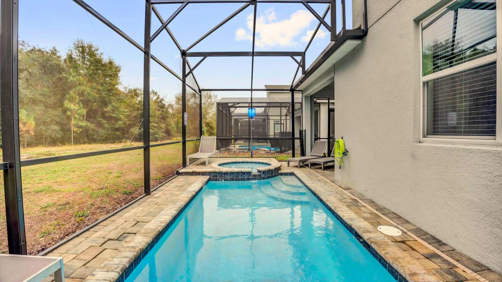 A narrow private pool and hot tub are enclosed within a screened lanai, featuring light blue water and brick-patterned coping. Beyond the screen, a grassy area and lush trees provide a natural backdrop under a partly cloudy sky.