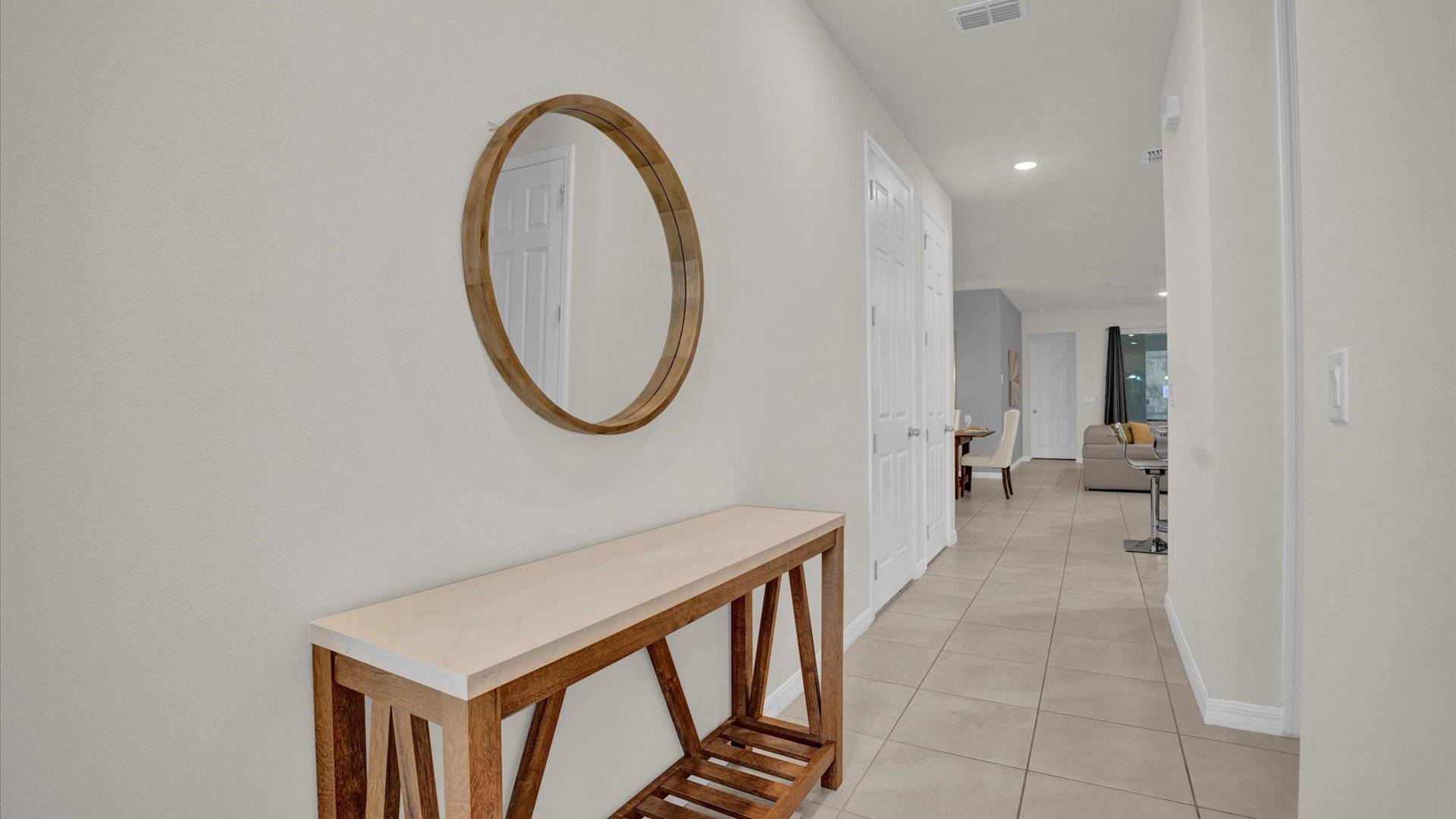A bright foyer features light beige walls, a large round wooden-framed mirror, and a rustic wooden console table with a light-colored top. The floor is tiled in a neutral tone, leading into a hallway with white doors.
