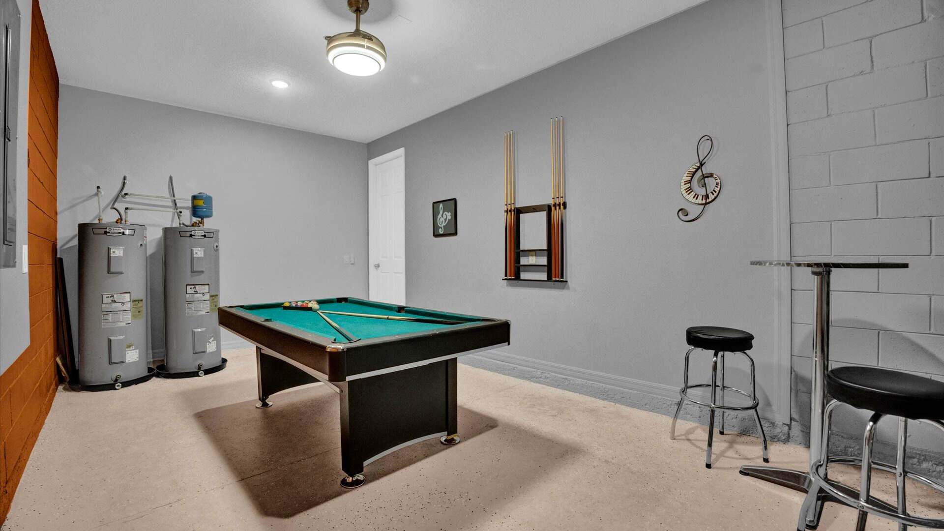 A game room featuring a green-felt pool table, two large water heaters, and a high-top table with two bar stools, all set against light gray walls and a concrete floor. A ceiling fan and a music-themed wall decor are also visible.