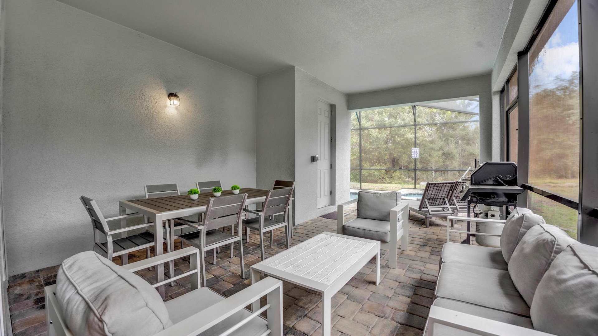 Covered patio area with a long outdoor dining table and chairs, a white outdoor lounge set, and a BBQ grill, all on brick pavers, with a view towards the screened pool area and natural landscape.