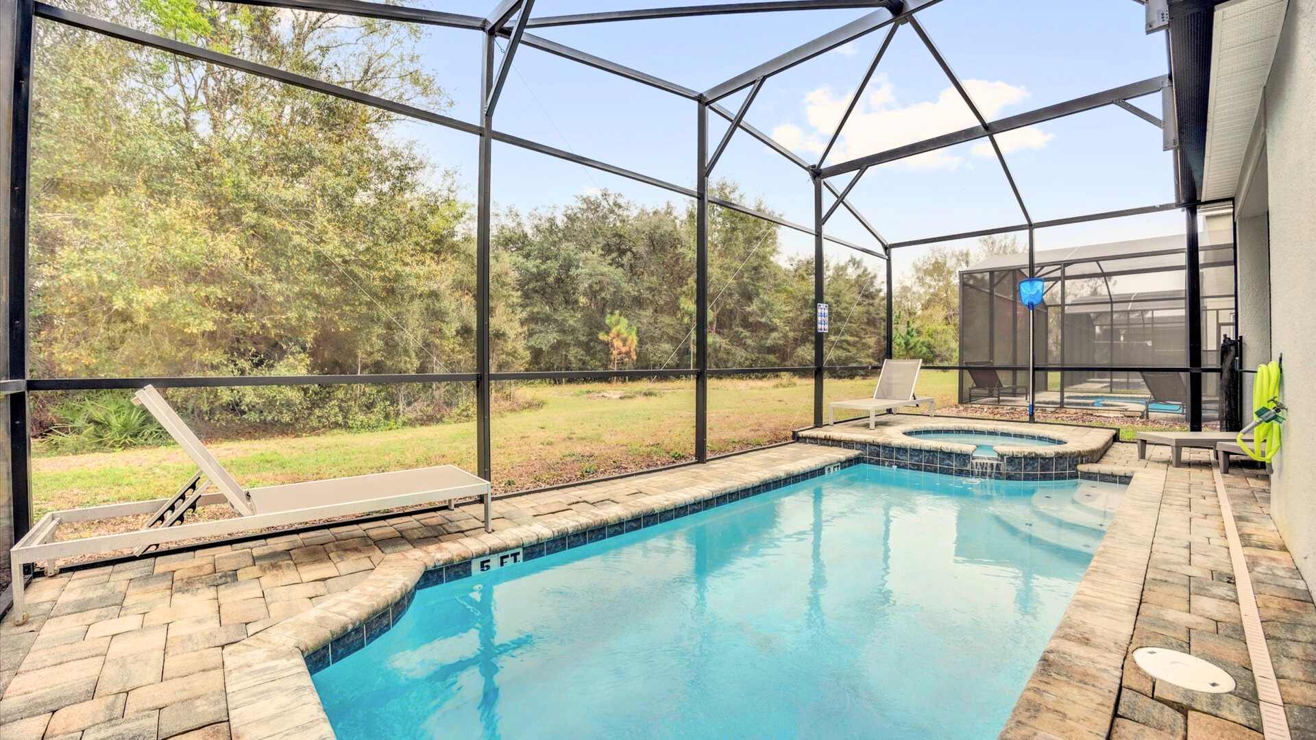 Expansive screened pool area showcasing a rectangular swimming pool with a shallow end and an integrated hot tub, surrounded by brick pavers and multiple lounge chairs, overlooking a natural landscape.