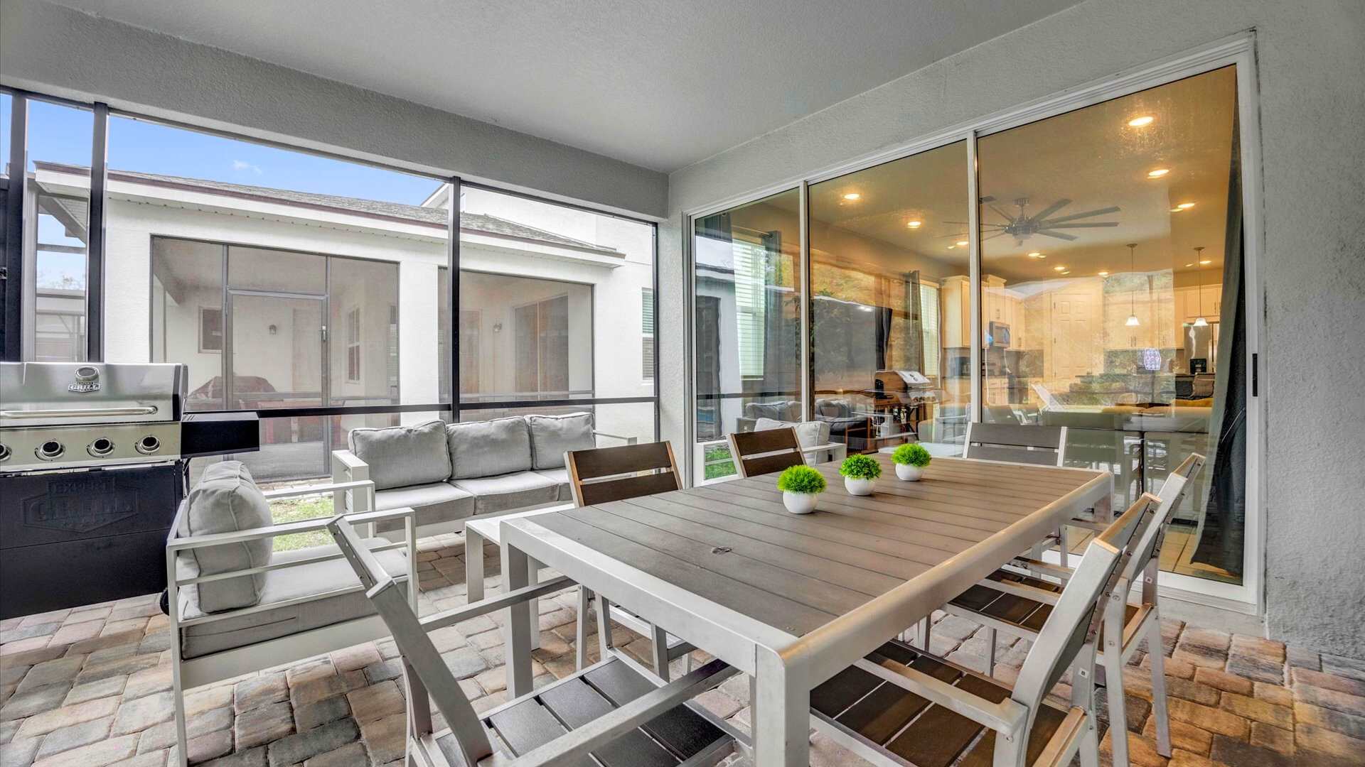 Spacious covered patio with a large outdoor dining table and chairs, a comfortable outdoor sofa and armchairs, and a BBQ grill, all on brick pavers within a screened enclosure.