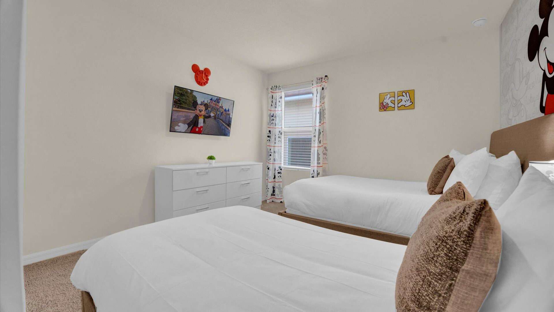 An alternate view of the Mickey Mouse-themed bedroom, showcasing a wall-mounted TV above a white dresser. The window is adorned with Mickey Mouse curtains, and one of the two beds with white bedding and brown accent pillows is in the foreground.