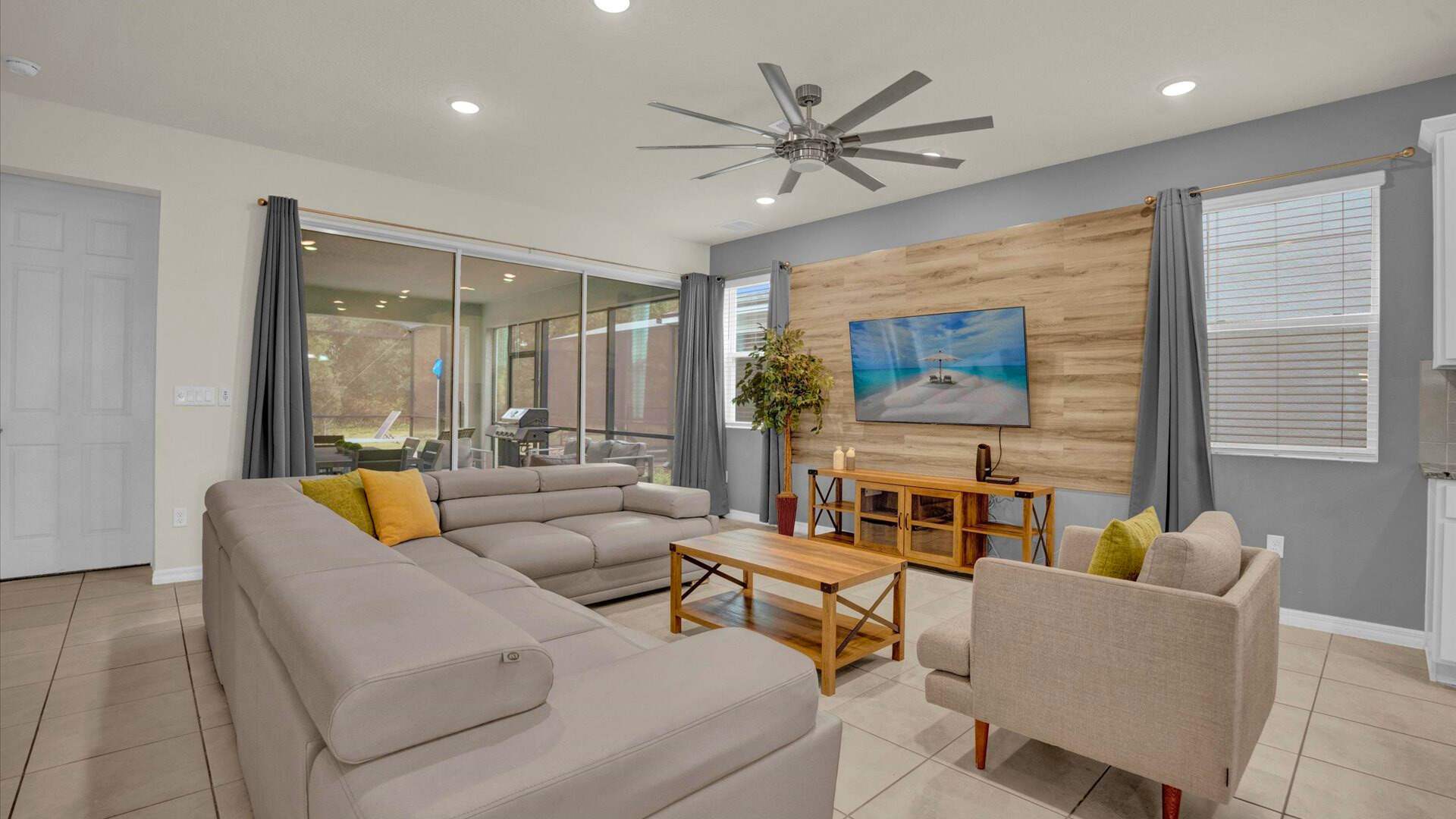 Bright living room with a large light gray sectional sofa and an armchair, centered around a wooden coffee table. A wall-mounted TV on a wood-paneled accent wall and a sliding glass door leading to an outdoor patio create a comfortable and connected space.