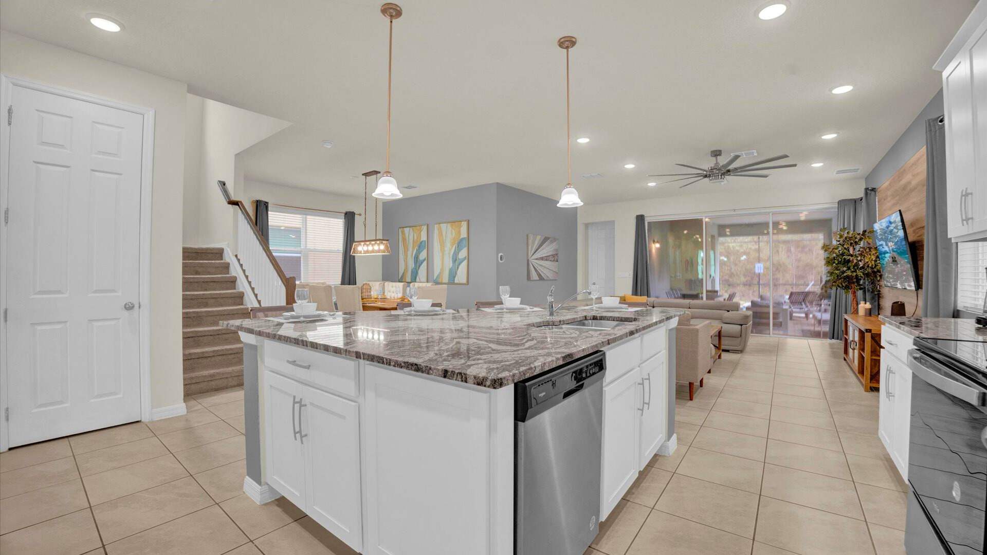 Bright and spacious kitchen centered around a large granite island with a double sink and dishwasher, offering ample counter space. White cabinetry, pendant lighting, and a view of the staircase complete this functional and inviting culinary area.
