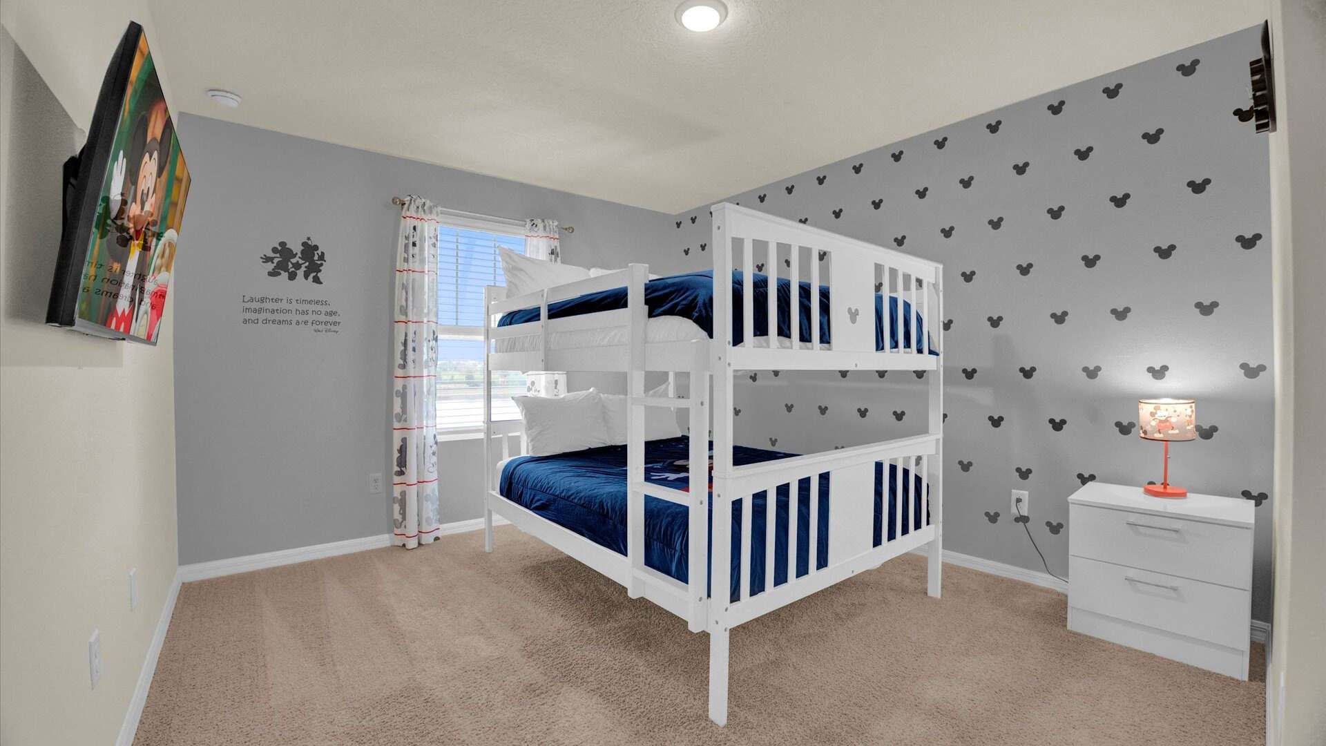 A fun, Mickey Mouse-themed bedroom featuring a white bunk bed with vibrant blue bedding. The grey walls are decorated with black Mickey Mouse head silhouettes and a motivational quote, complemented by Mickey Mouse curtains and a wall-mounted TV.