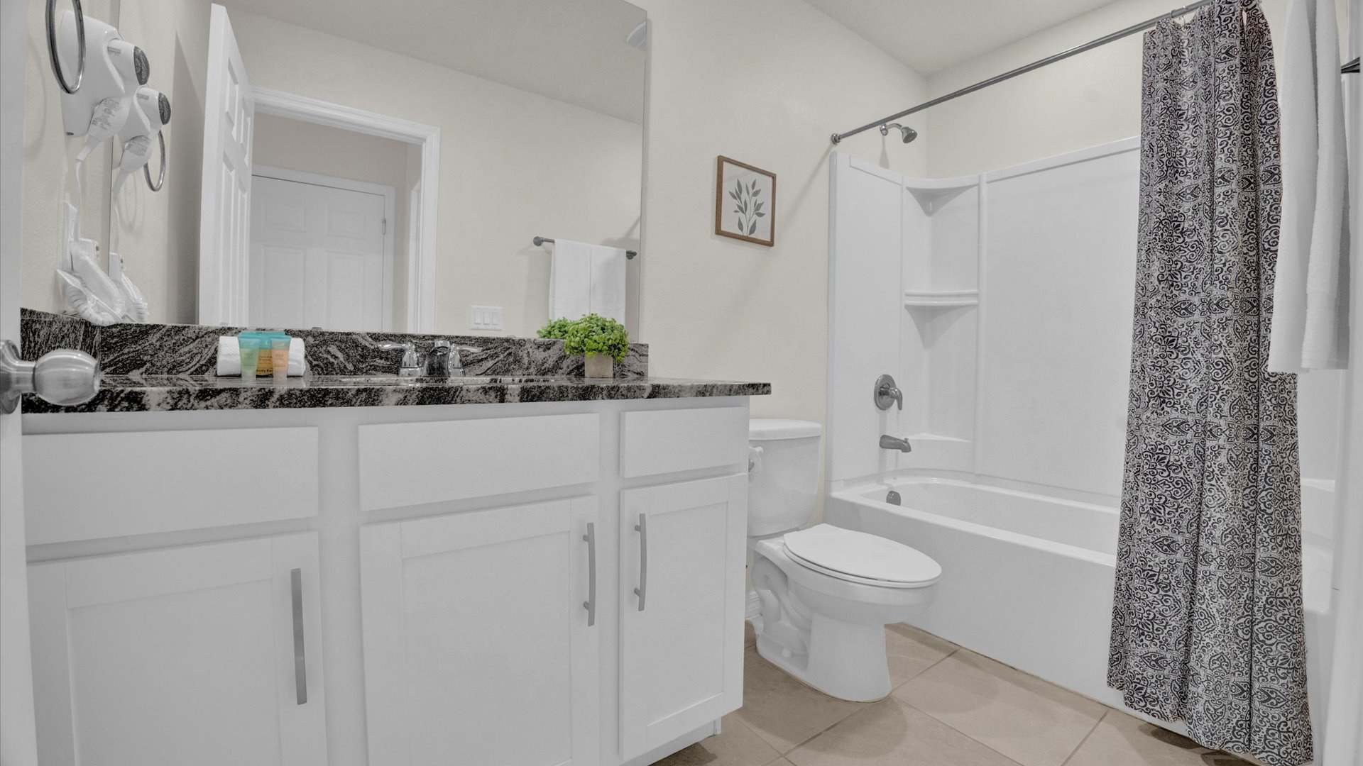 A clean bathroom featuring a white vanity with a dark granite countertop and a single sink, beneath a large mirror. A white toilet and a bathtub with a shower and a black and white patterned curtain are visible on the right, all set on a tiled floor.