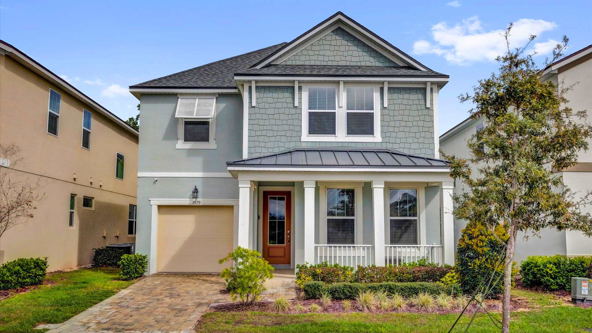 The front exterior of a two-story house with light blue siding and gray shingle accents, featuring a covered porch with white columns and railing. A paved driveway leads to a single-car garage, surrounded by manicured landscaping.
