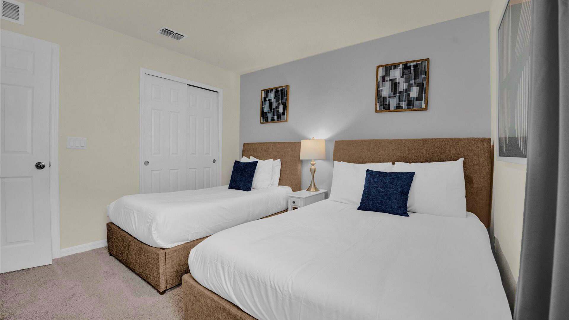 A bright bedroom featuring two beds with brown upholstered headboards, white bedding, and navy blue accent pillows. A light blue accent wall behind the beds is adorned with two abstract framed artworks, complementing the light yellow walls and white sliding closet doors.