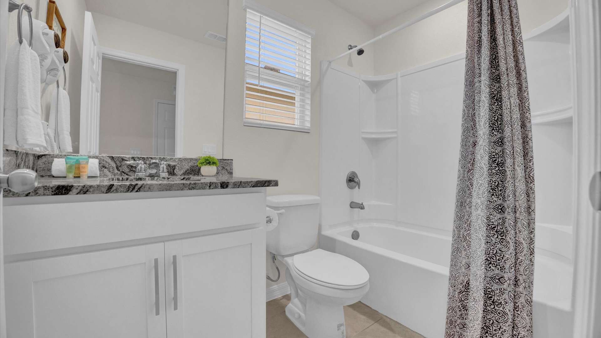 A bright bathroom featuring a white vanity with a dark granite countertop and a single sink, beneath a large mirror. A window is positioned above the toilet, next to a bathtub with a shower and a black and white patterned curtain, all on a tiled floor.