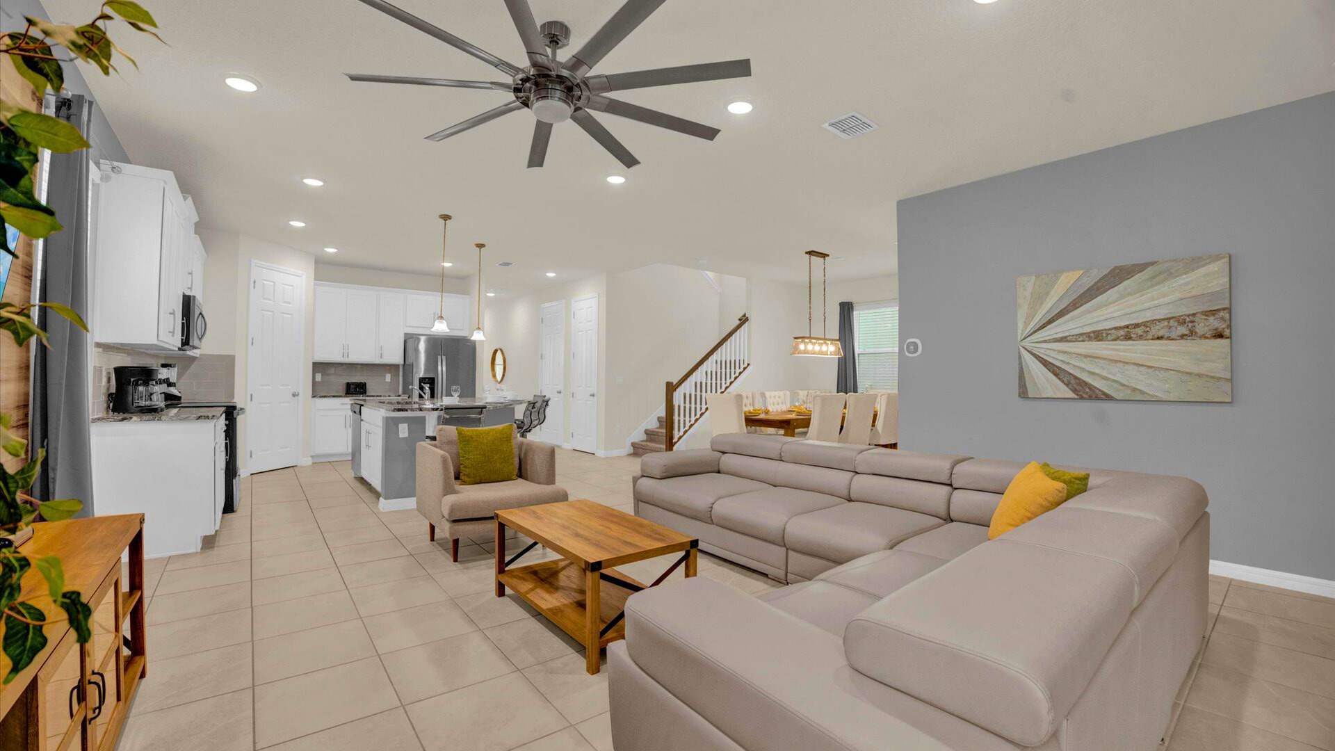 Expansive open-concept living area featuring a large light gray sectional sofa and a wooden coffee table, with a view towards the modern kitchen and dining room. A grand ceiling fan and light gray walls create a bright, inviting atmosphere.