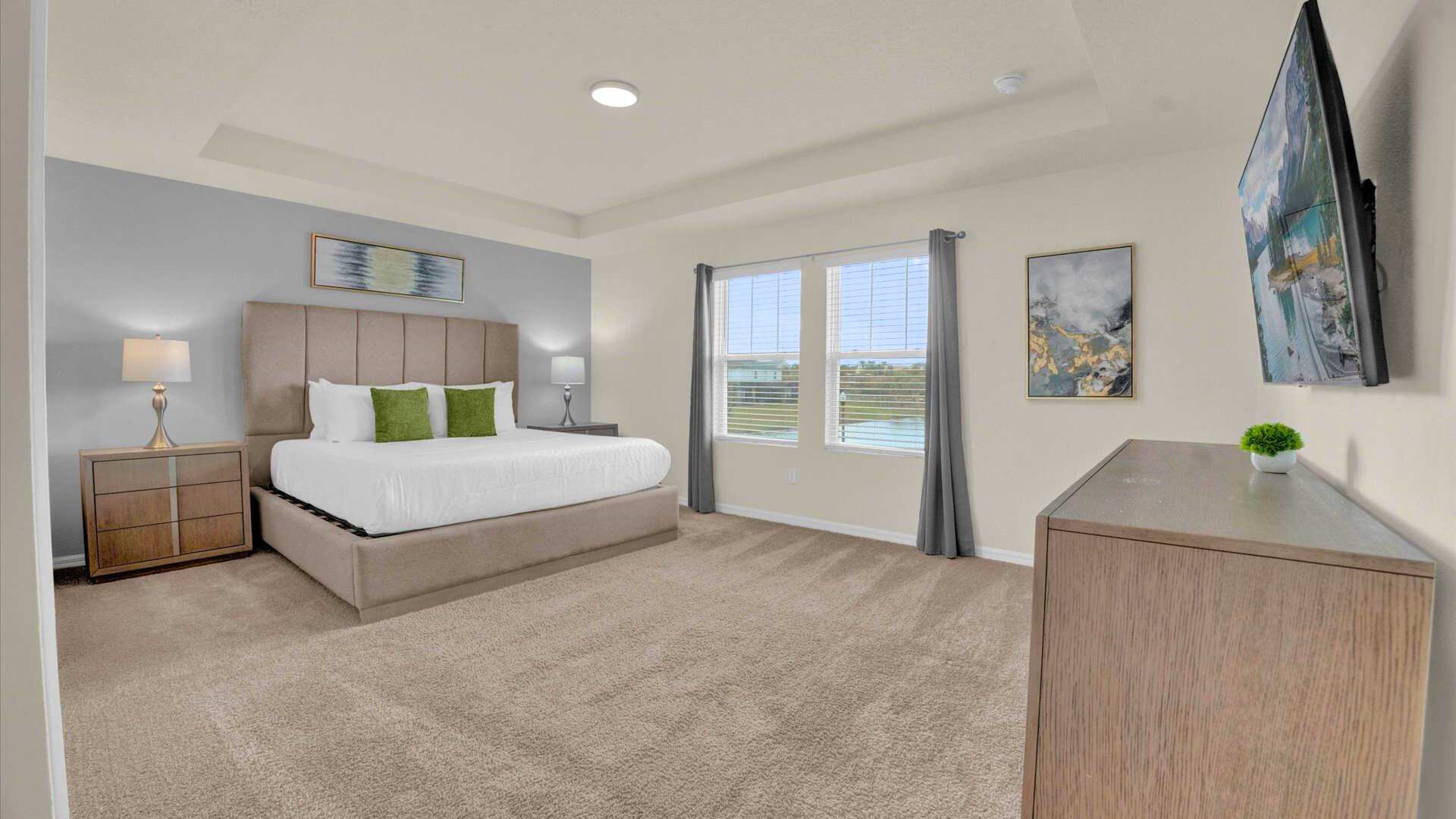 Master bedroom with light beige carpet, a king-sized bed with a tall upholstered headboard, and two nightstands with lamps. A light grey accent wall behind the bed complements the room's neutral tones, with windows offering a lake view.