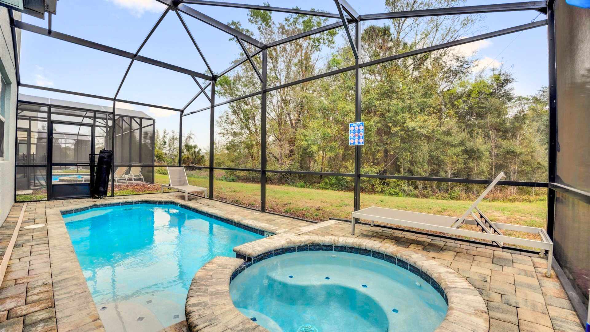 Screened outdoor area featuring a rectangular swimming pool and an adjacent circular hot tub, both surrounded by brick pavers, with lounge chairs and a view of lush greenery beyond the screen.