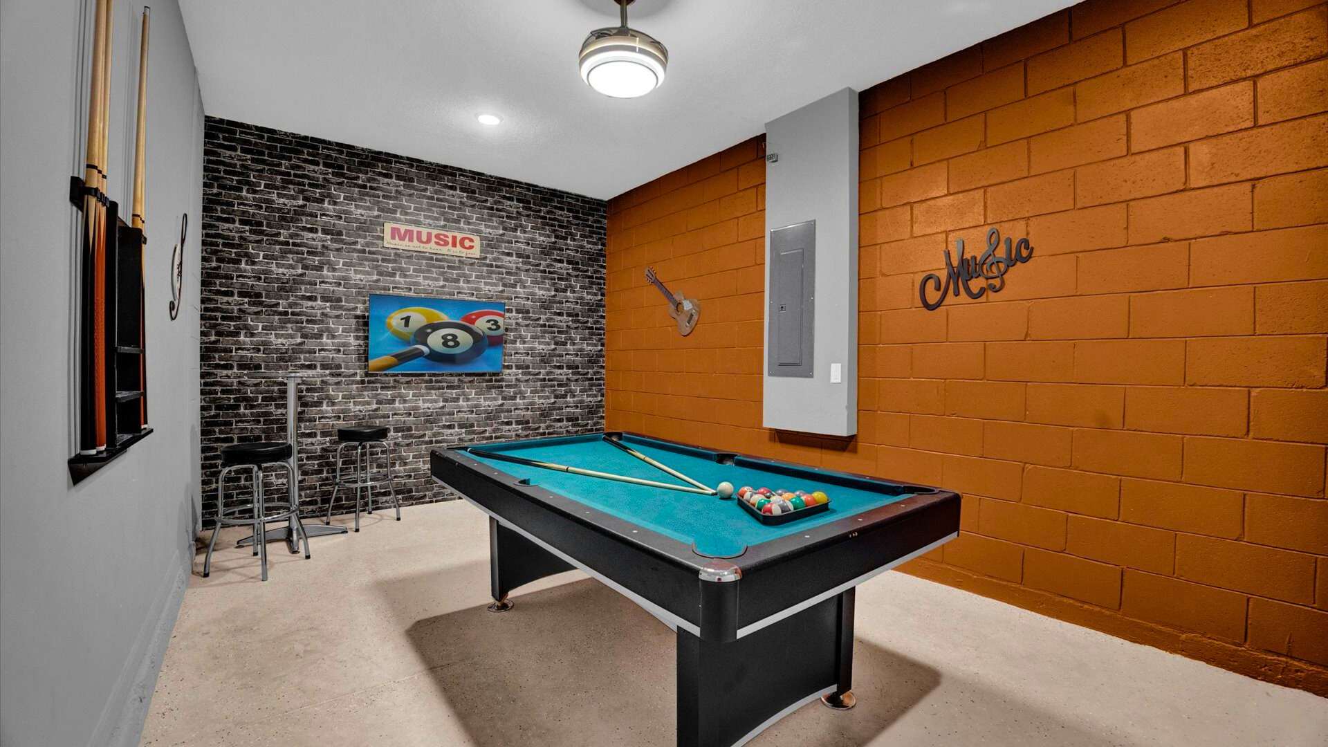 Game room featuring a teal-felt pool table with billiard balls, a black brick accent wall with a 'MUSIC' sign and pool cue rack, and a textured orange wall, under a modern ceiling light.