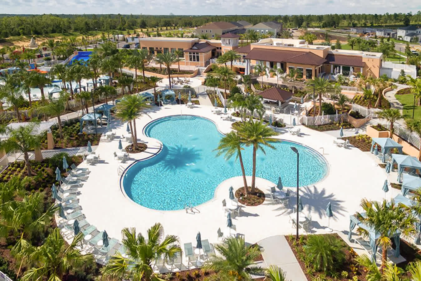 An aerial shot captures a large, irregularly shaped communal pool with vibrant blue water, surrounded by a spacious deck filled with lounge chairs and umbrellas. Palm trees and lush landscaping enhance the resort-like atmosphere.