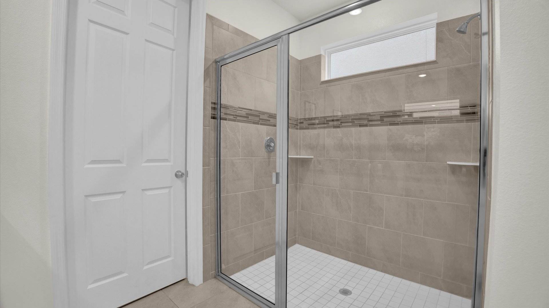 Walk-in shower with light brown and grey tiled walls, a decorative horizontal tile accent, and a clear glass enclosure. A small frosted window above the shower provides natural light, and the floor is white mosaic tile.