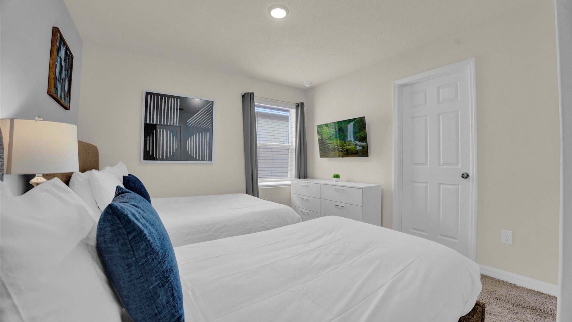 View of bedroom-4 showing a white dresser with a wall-mounted TV above it, positioned between two brown upholstered beds. The room has a window with blinds and a light-colored wall.