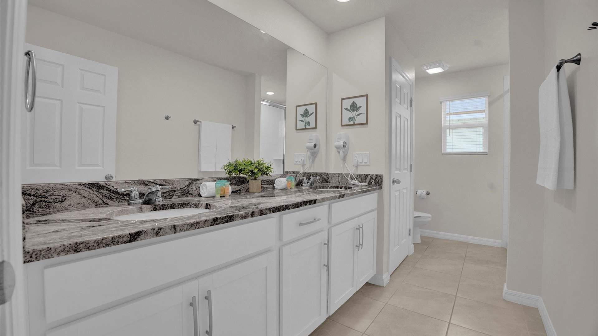 A spacious bathroom with a double vanity featuring a dark granite countertop, white cabinets, and two sinks. A large mirror spans the width of the vanity, and two framed botanical prints adorn the wall.