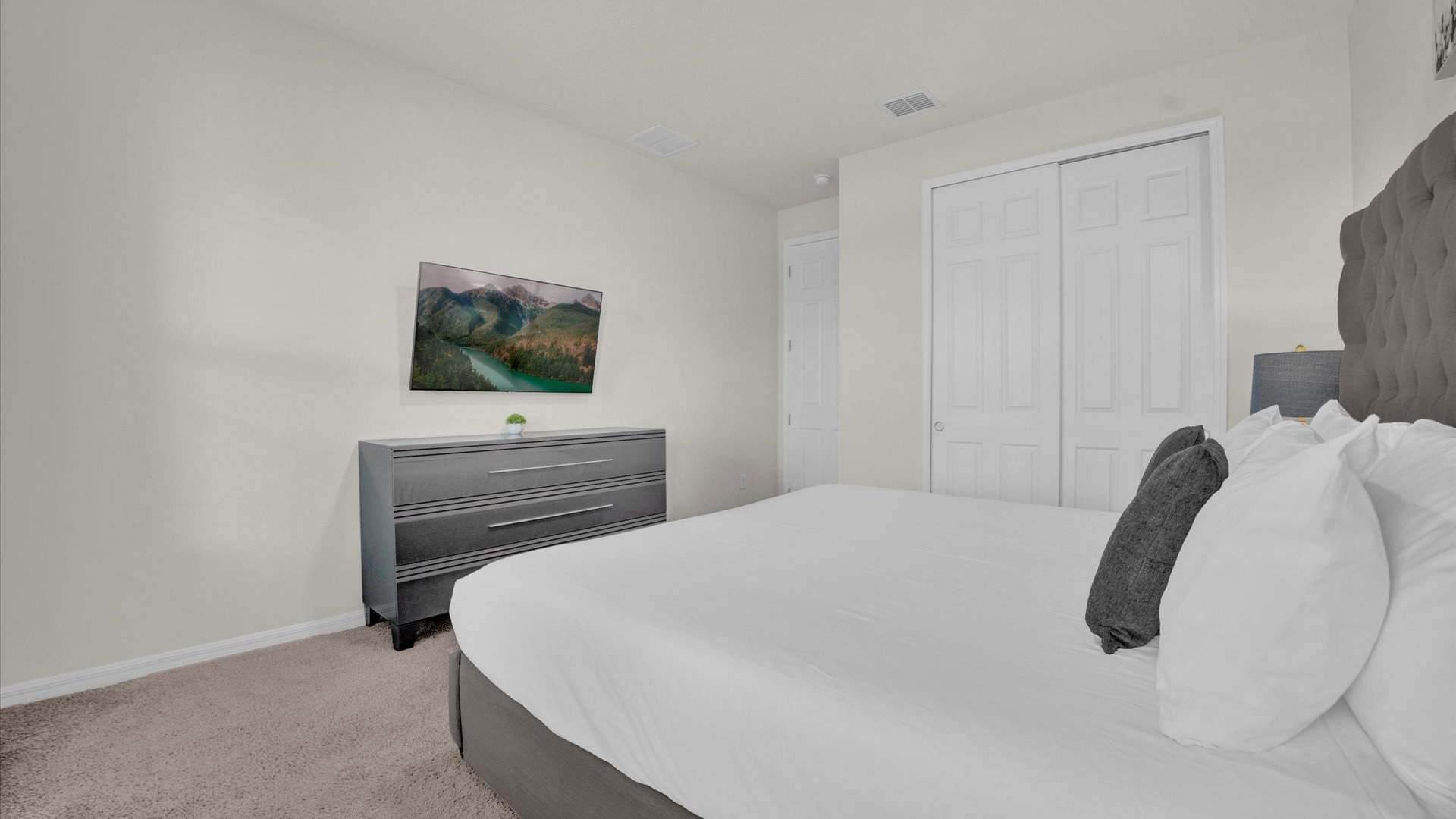 View of bedroom-2 showing a dark gray dresser with a wall-mounted TV above it, and a large white sliding closet door. The gray upholstered king bed is partially visible.