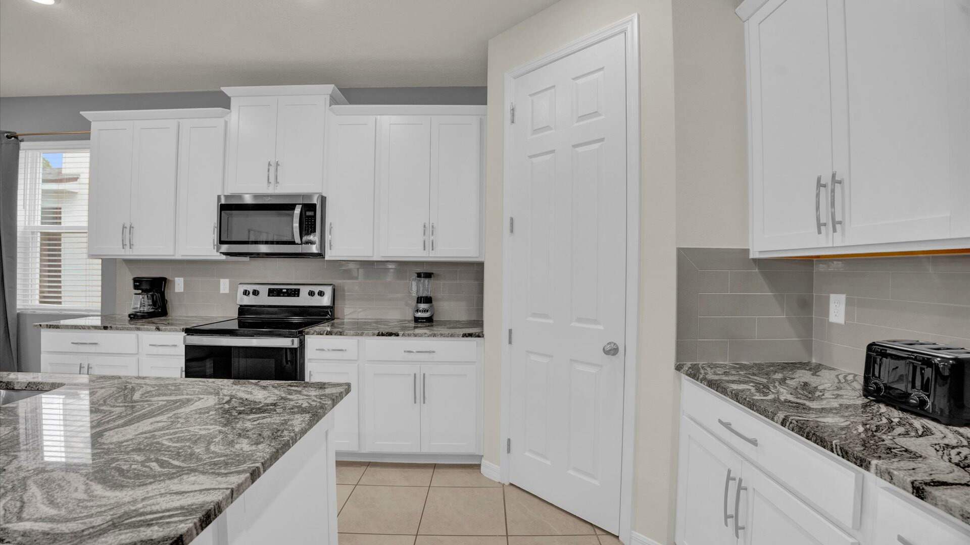 Modern kitchen featuring white shaker cabinets, dark granite countertops with grey veining, a grey subway tile backsplash, and stainless steel appliances including a microwave and electric stove. The floor is tiled in a neutral beige.