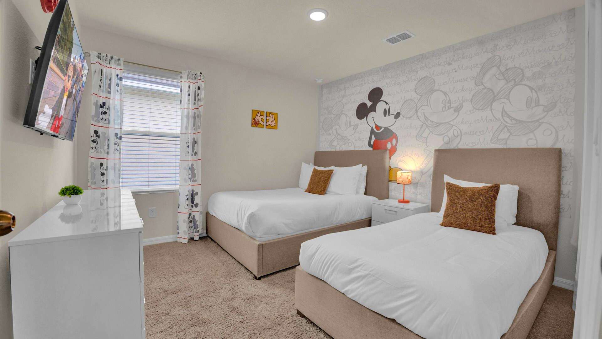A vibrant Mickey Mouse-themed bedroom featuring two beds with light brown upholstered headboards, white bedding, and brown accent pillows. A large Mickey Mouse graphic adorns the accent wall, complemented by Mickey Mouse curtains and a wall-mounted TV.