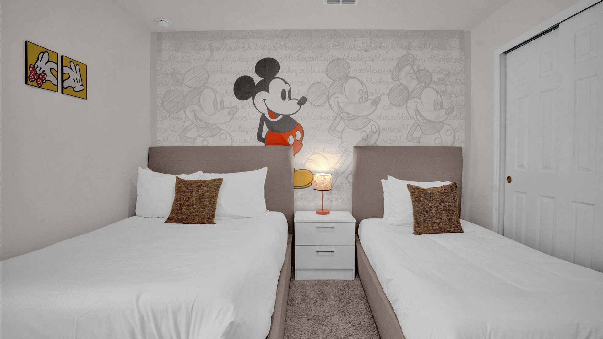 A close-up view of the Mickey Mouse-themed bedroom, highlighting the two beds with light brown upholstered headboards and the prominent Mickey Mouse graphic on the accent wall. A white nightstand with a lamp sits between the beds, and white sliding closet doors are visible.