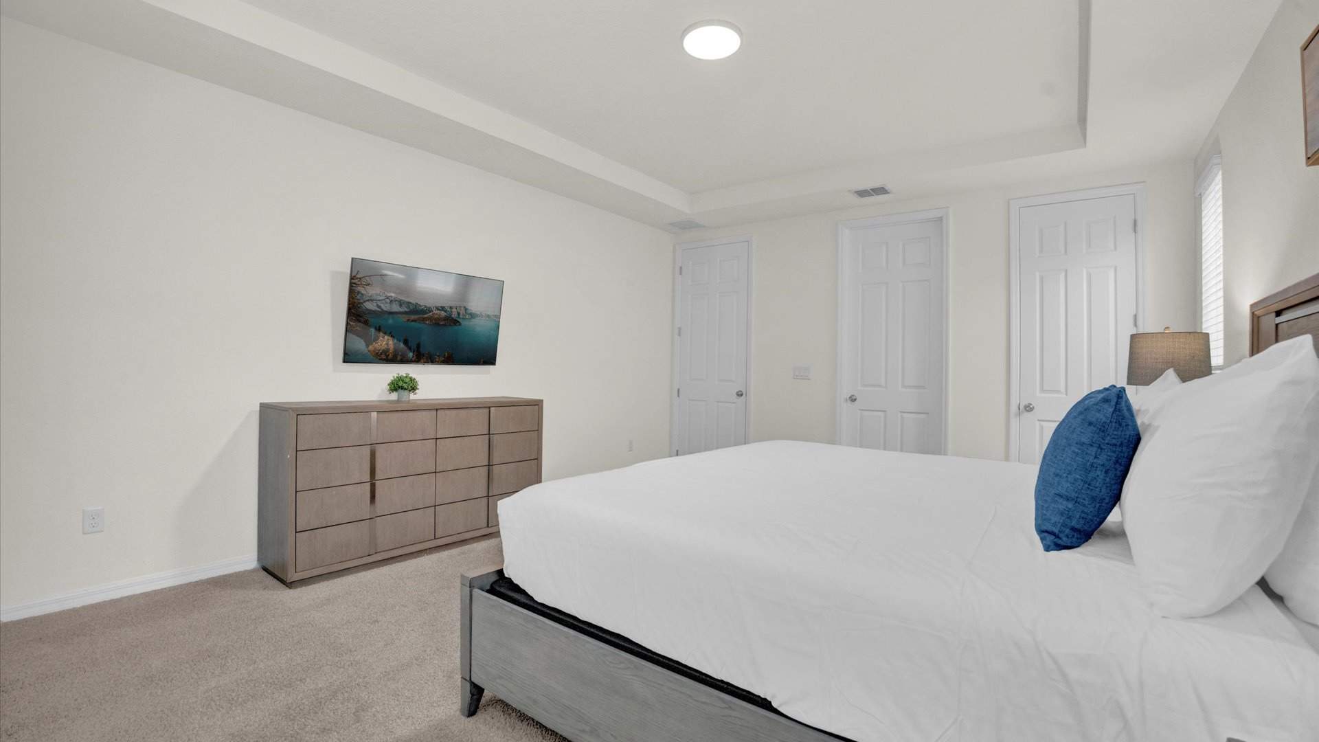 View of the bedroom showing a long wooden dresser with a wall-mounted TV above it. The room has light beige carpet, a tray ceiling with recessed lighting, and two white doors, likely leading to a closet and bathroom.