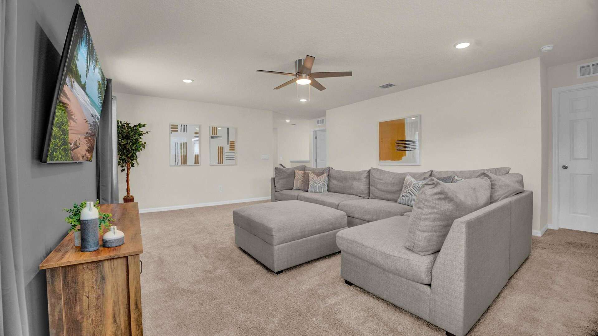 Bright living room with light beige carpet, a large L-shaped grey sectional sofa, and a wooden TV console with a wall-mounted TV. Decorative wall art and a potted plant add accents to the space, illuminated by recessed lighting and a ceiling fan.