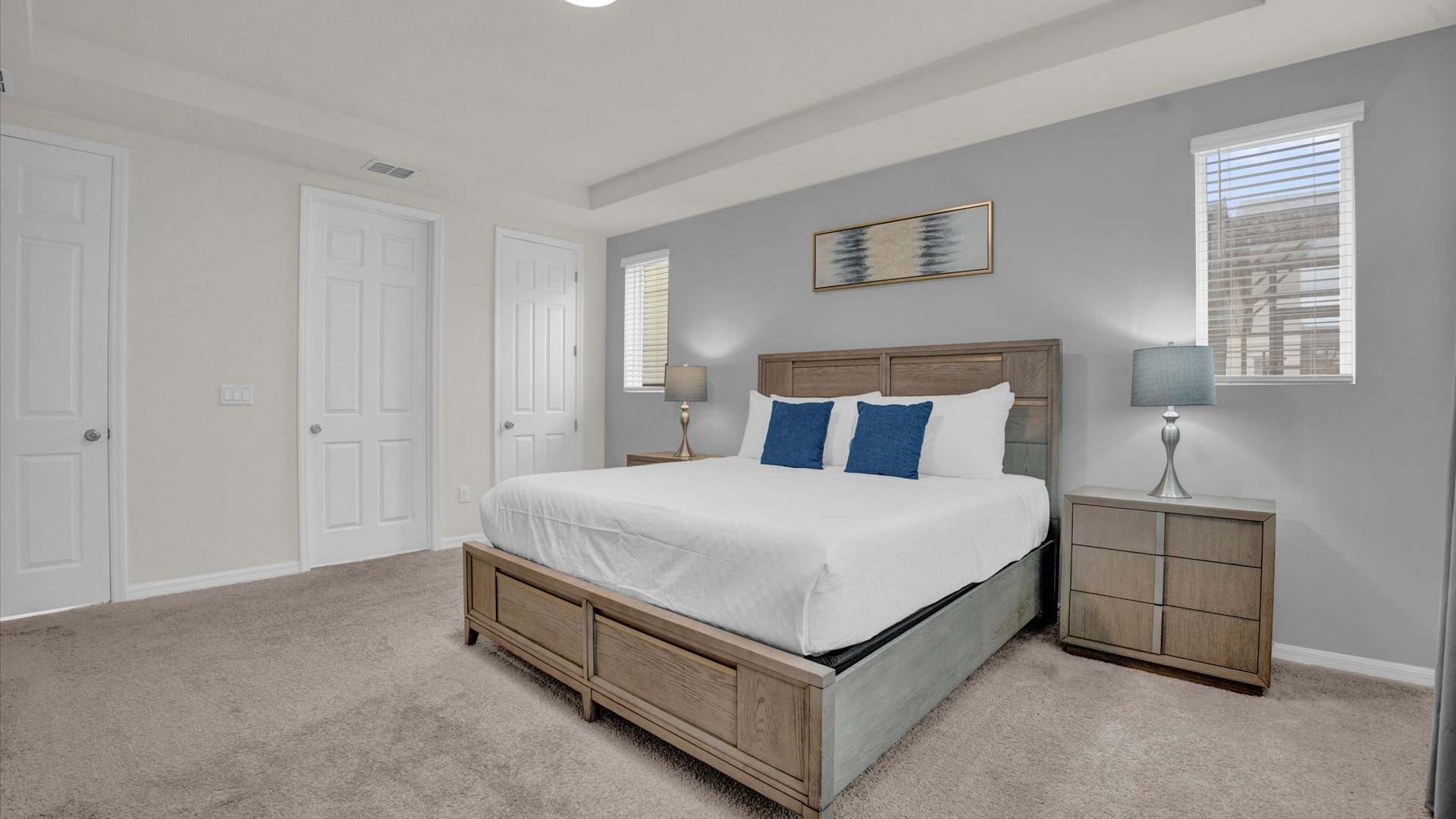 A spacious bedroom featuring a king-sized bed with a wooden frame and headboard, white bedding, and two blue accent pillows. Two nightstands with lamps flank the bed, set against a light gray accent wall with abstract art above the headboard.