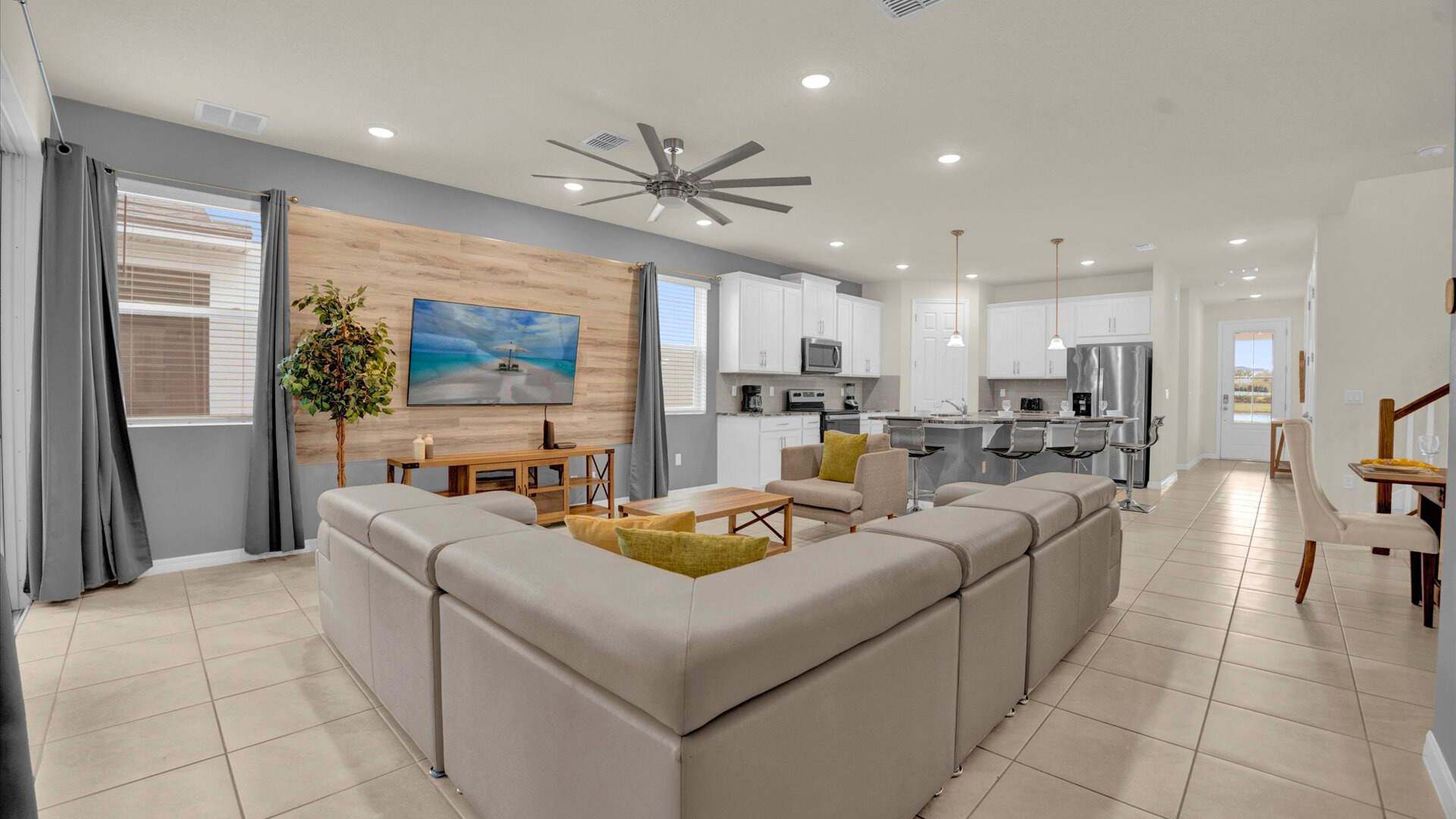 Spacious open-concept living room with a large light gray sectional sofa and an armchair, facing a wall-mounted TV above a wooden console. A striking wood-paneled accent wall and a large ceiling fan enhance the modern aesthetic.