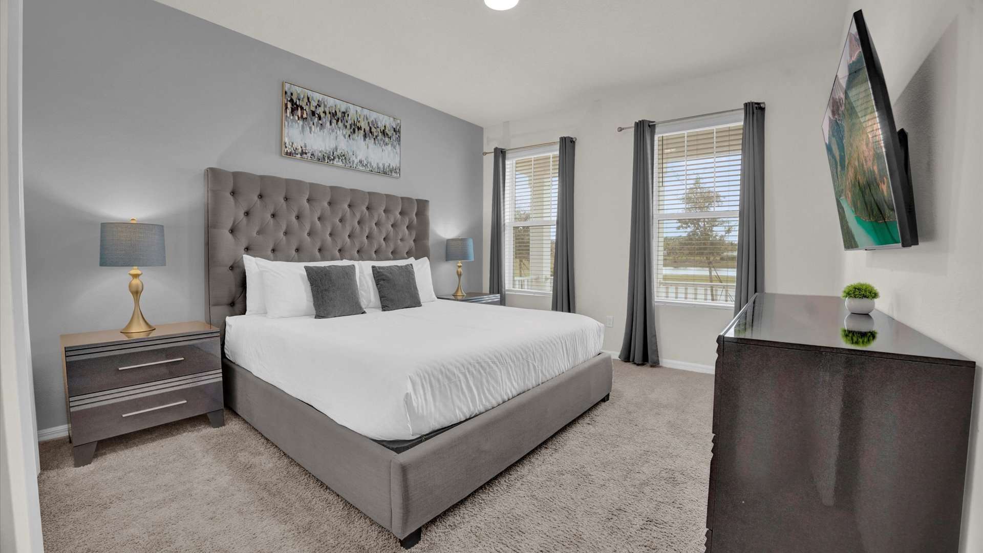 A stylish bedroom with a gray upholstered king bed featuring a tall tufted headboard, white bedding, and dark gray accent pillows. Two modern nightstands with lamps flank the bed, and a wall-mounted TV is opposite.