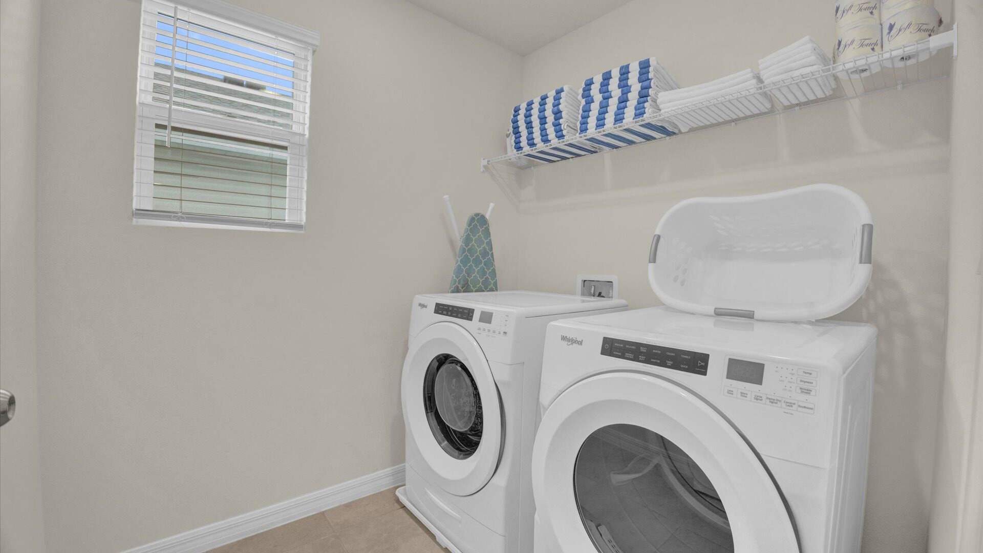 A compact laundry room features a white front-loading washer and dryer set, with a window providing natural light. A wire shelf above holds neatly folded towels, and an ironing board leans against the wall.