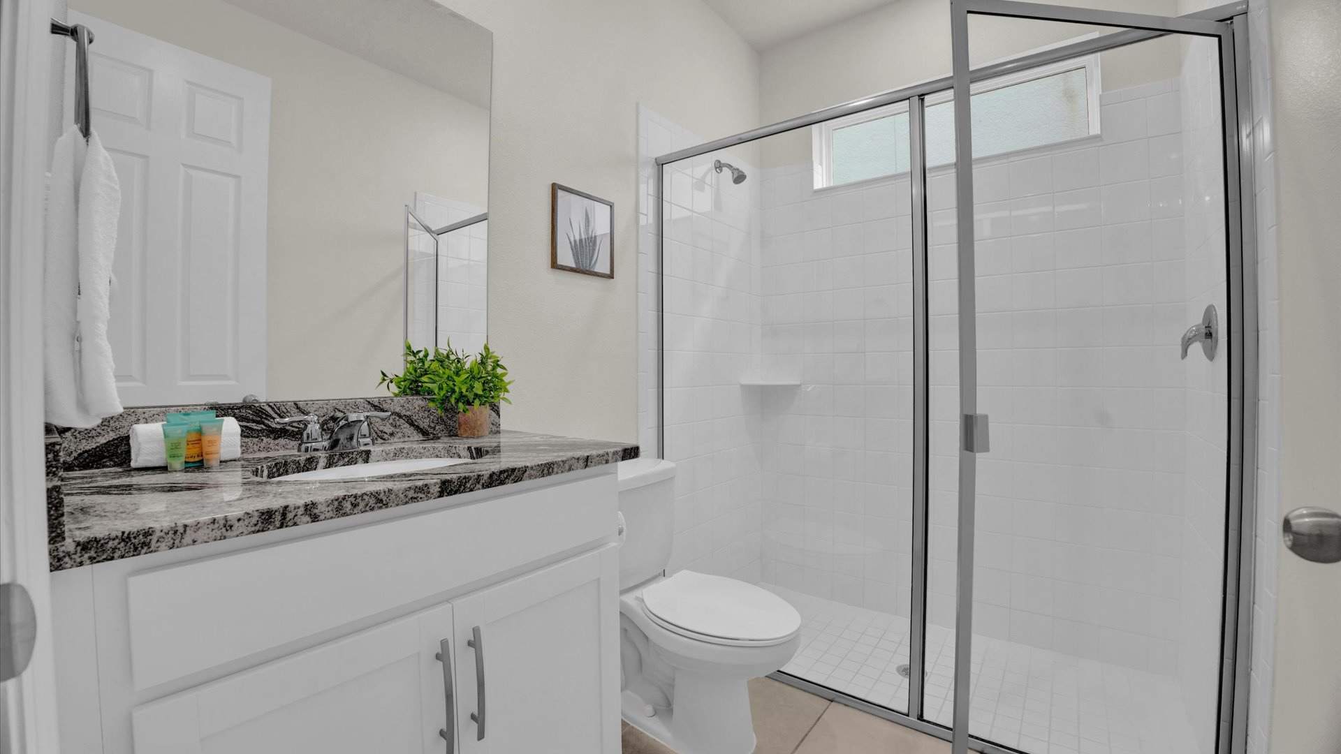 A modern bathroom featuring a white vanity with a dark granite countertop and a single sink, beneath a large mirror. A spacious walk-in shower with a glass enclosure and white subway tiles is visible, alongside a white toilet, all on a tiled floor.