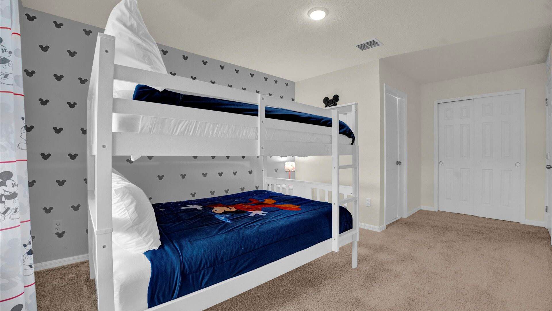 An alternate view of the Mickey Mouse-themed bunk bed room, showcasing the white bunk bed with blue bedding and a character blanket. The grey accent wall is patterned with black Mickey Mouse heads, and white closet doors are visible in the background.