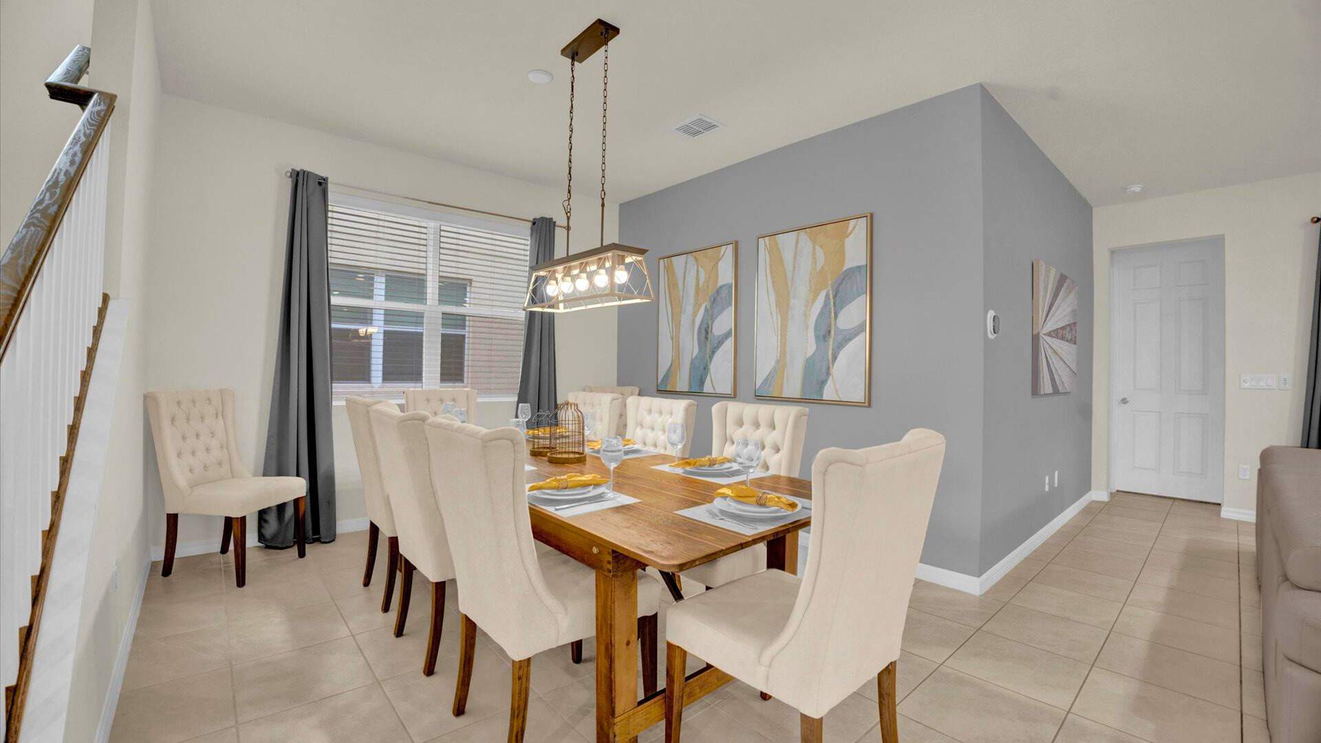 Elegant dining room with a long wooden table set for eight, surrounded by upholstered cream chairs, under a linear chandelier, with a gray accent wall adorned with two abstract art pieces.