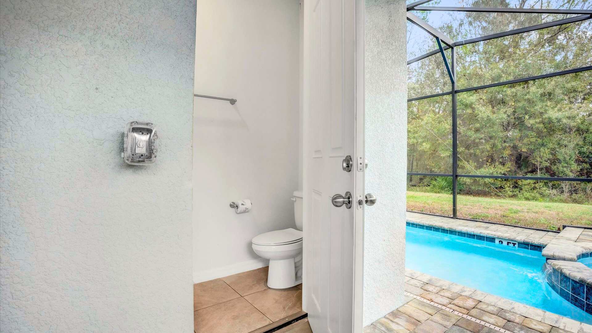 A small, functional bathroom with light-colored walls and neutral tiled flooring, featuring a white toilet and a towel bar. The open door reveals the private pool area outside, suggesting it's a convenient pool bath.