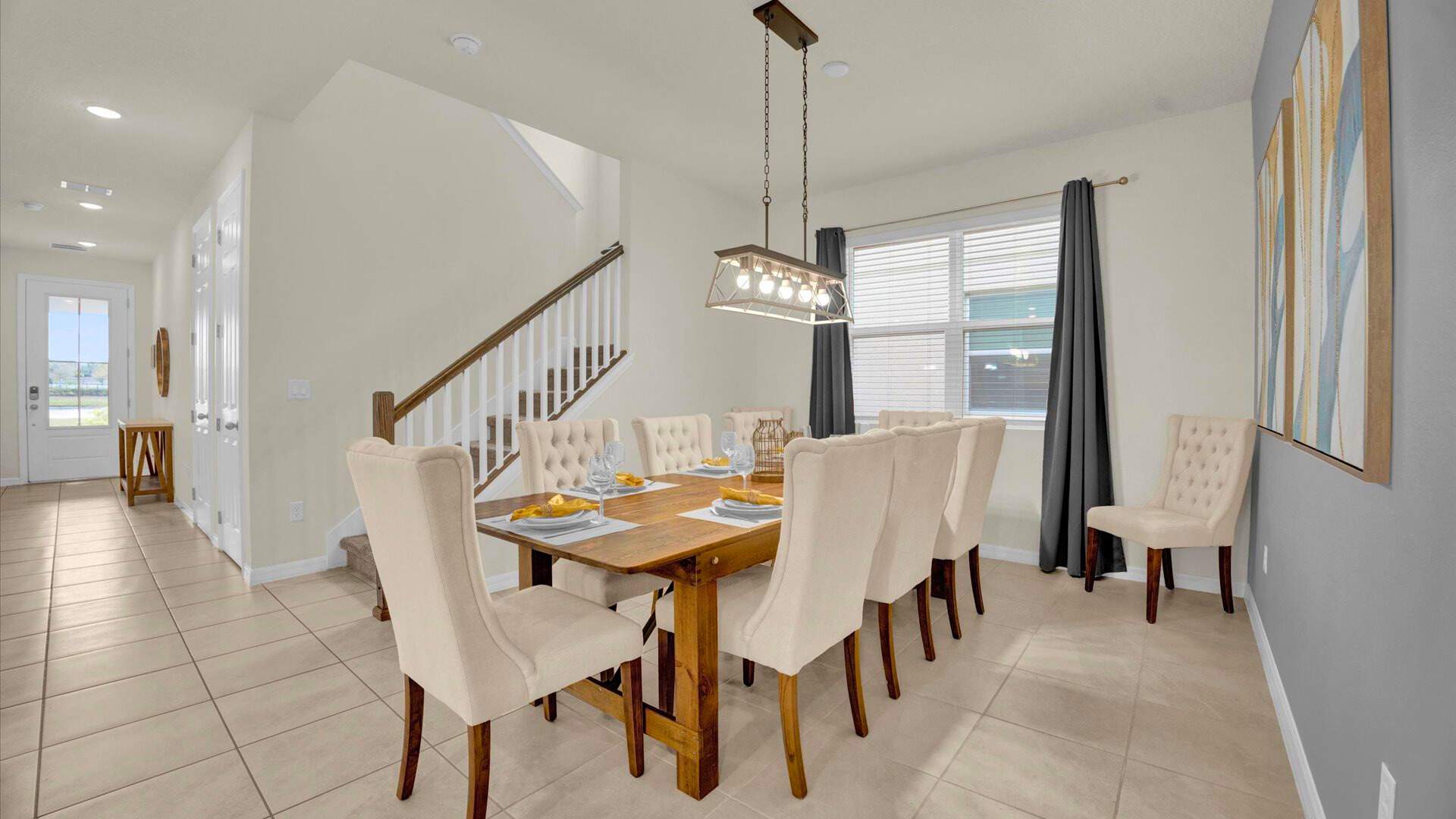 Elegant dining room featuring a long wooden table with eight upholstered chairs, set beneath a modern linear chandelier. A staircase with white balusters is visible in the background, adding to the home's spacious feel.